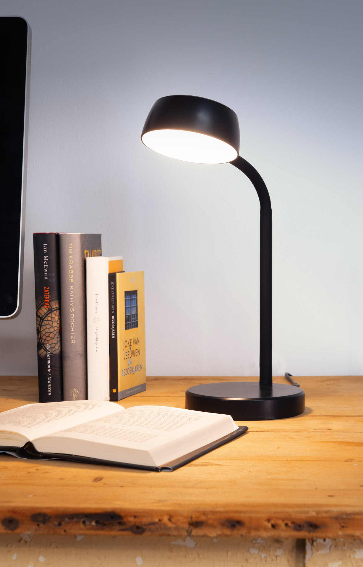 Eine schwarze Schreibtischlampe beleuchtet ein offenes Buch auf einem Holztisch. Links stehen Bücher. Ein Computerbildschirm ist teilweise sichtbar.