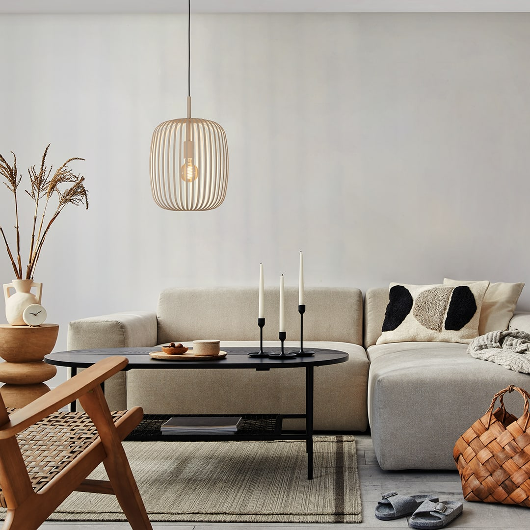 Een moderne woonkamer met een beige bank, zwarte salontafel, kaarsen en een hanglamp. Een gevlochten mand en slippers liggen op de vloer.