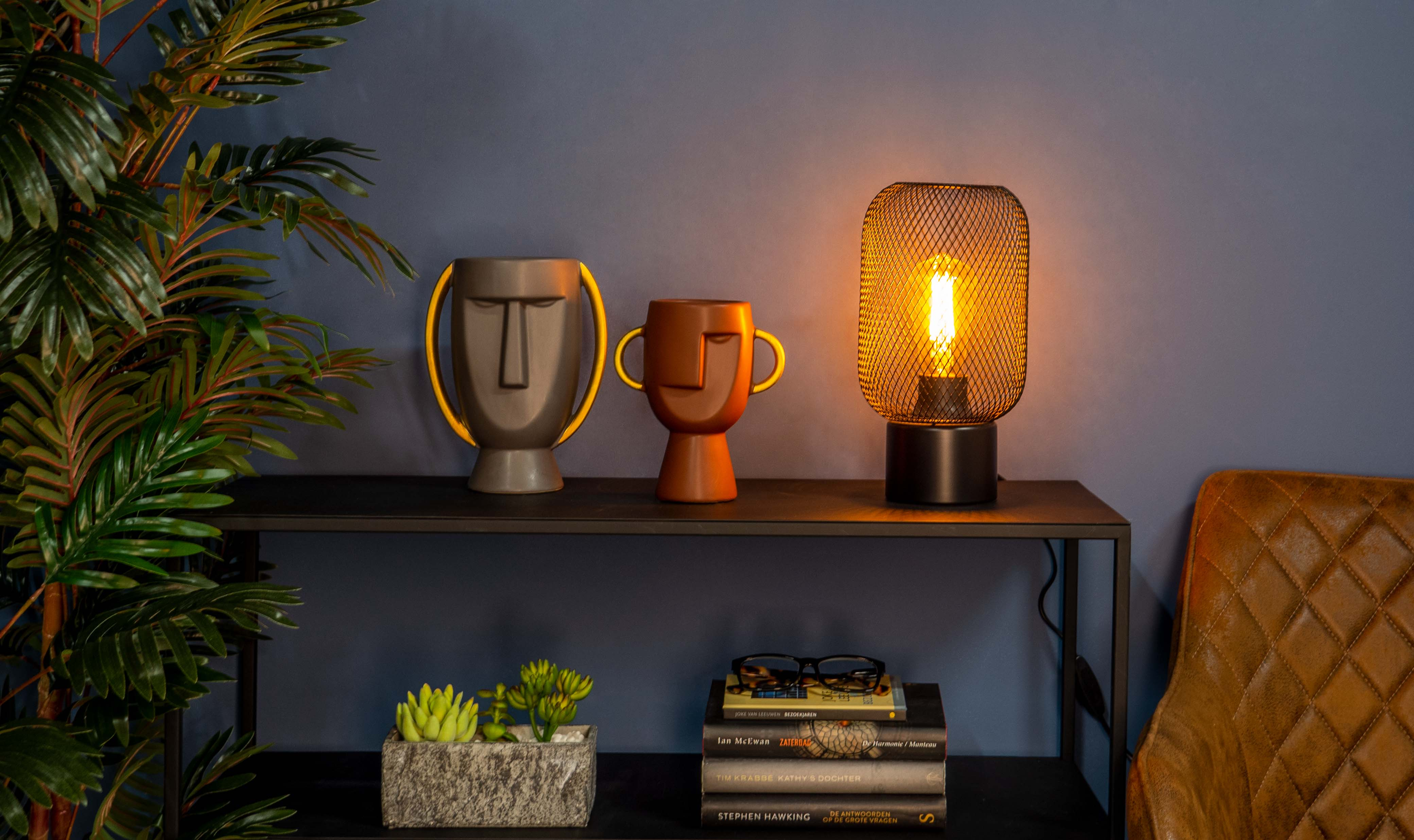 Ein Regal mit Dekoration: Vasen, Lampe, Bücher und Pflanzen. Ein Lederstuhl und Palmblätter im Hintergrund.