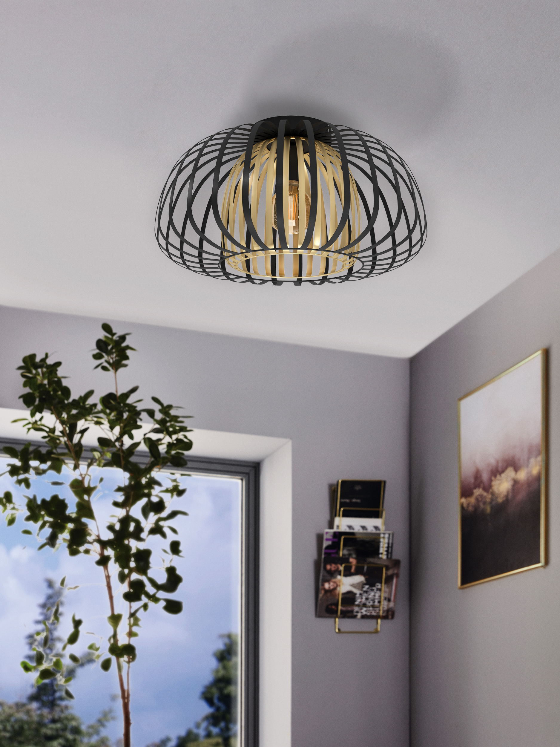 Eine moderne Deckenlampe, schwarzer Rahmen, goldfarbenes Innenleben und eine brennende Glühbirne. Neben einem Fenster mit Pflanze und Wand mit Bild.