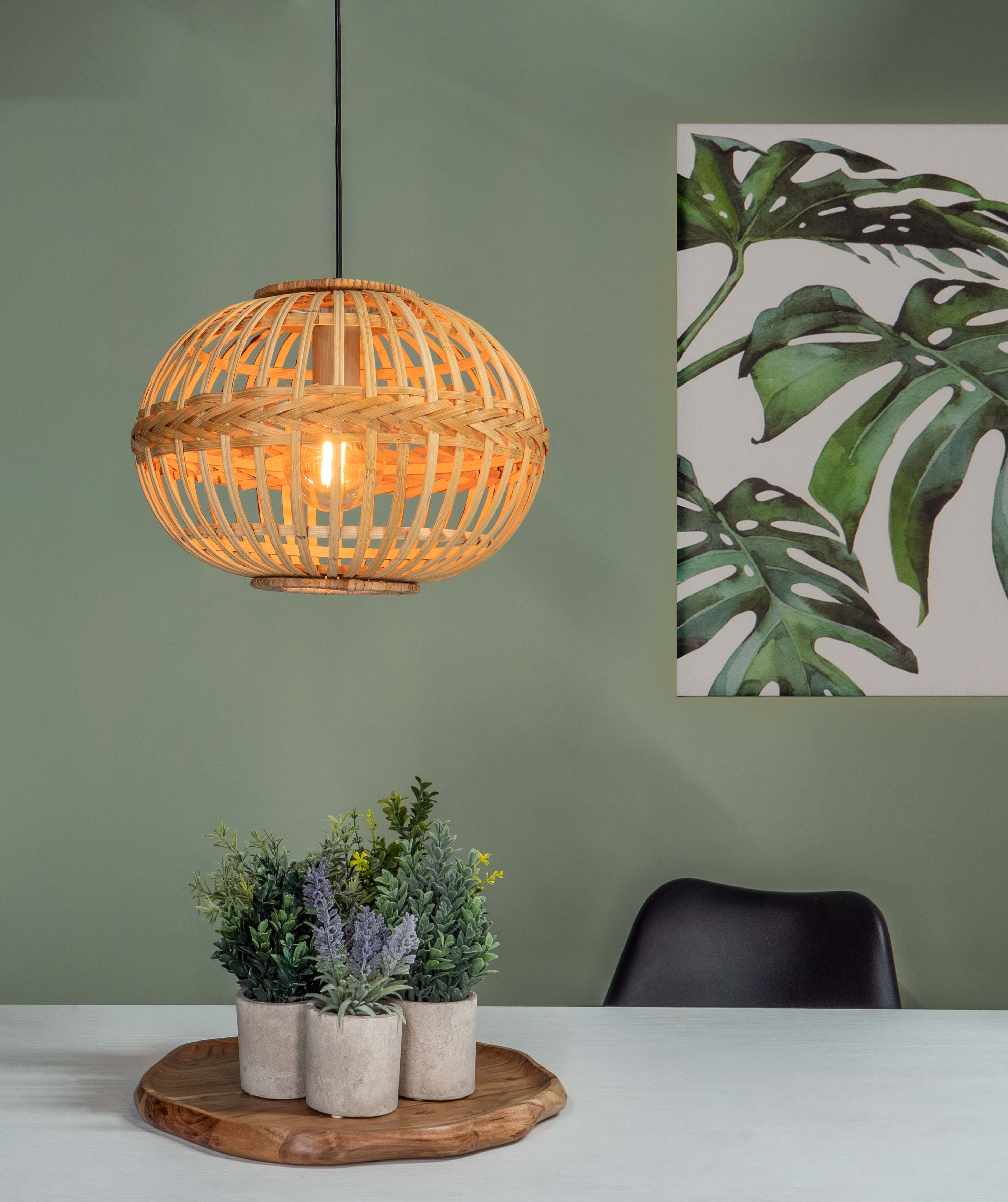 Tkana oprawa oświetleniowa wisi nad roślinami na drewnianej tacy. Na ścianie znajduje się grafika z liści monstery.