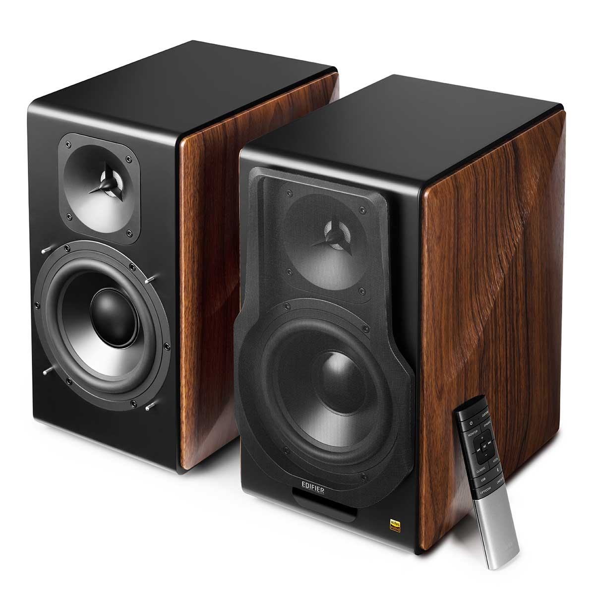Dwa czarne głośniki z bokami z fakturą drewna. Obok jednego leży pilot. Głośniki mają przetworniki pokryte siatką.