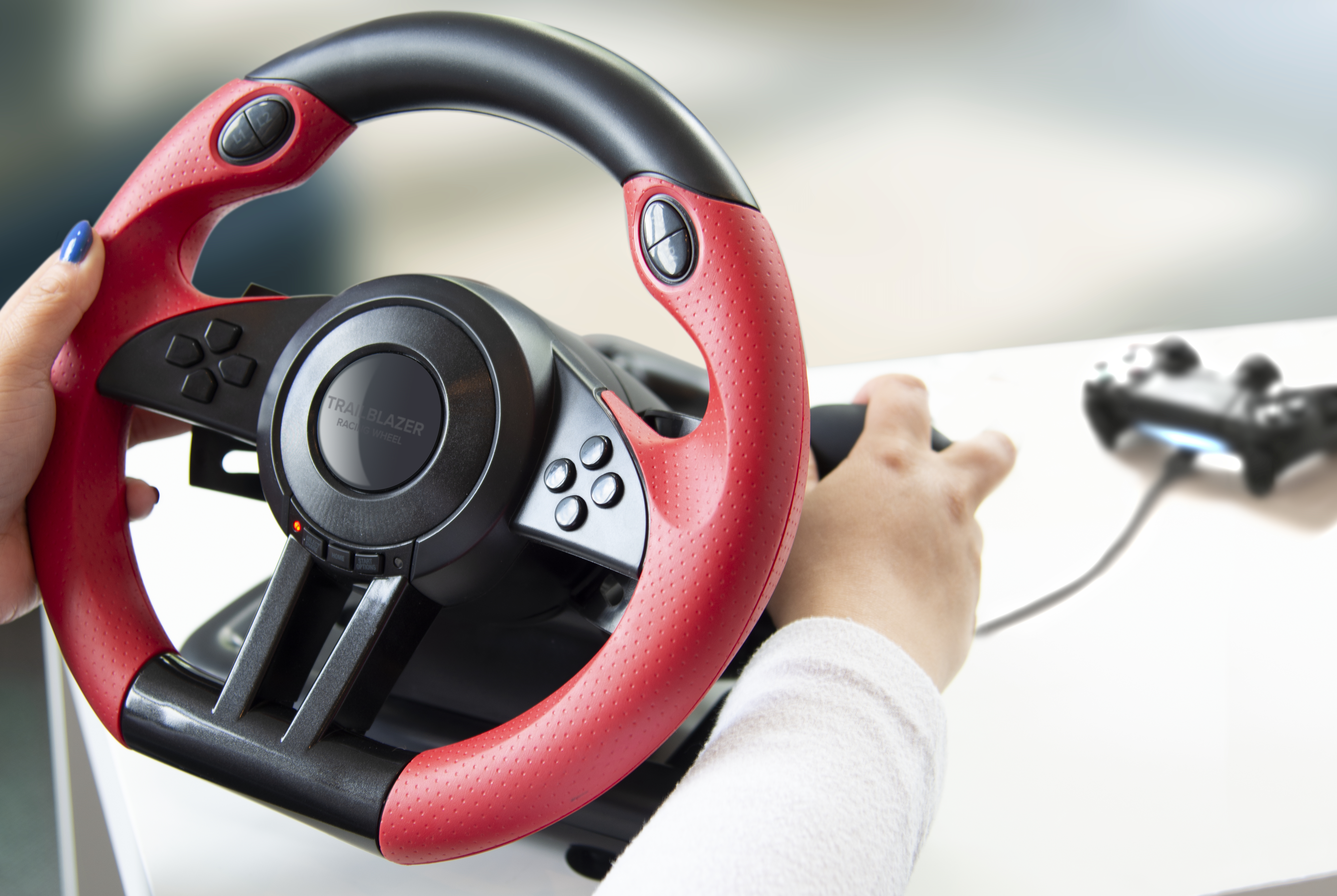 A person is holding a red and black gaming steering wheel with a controller in the background.