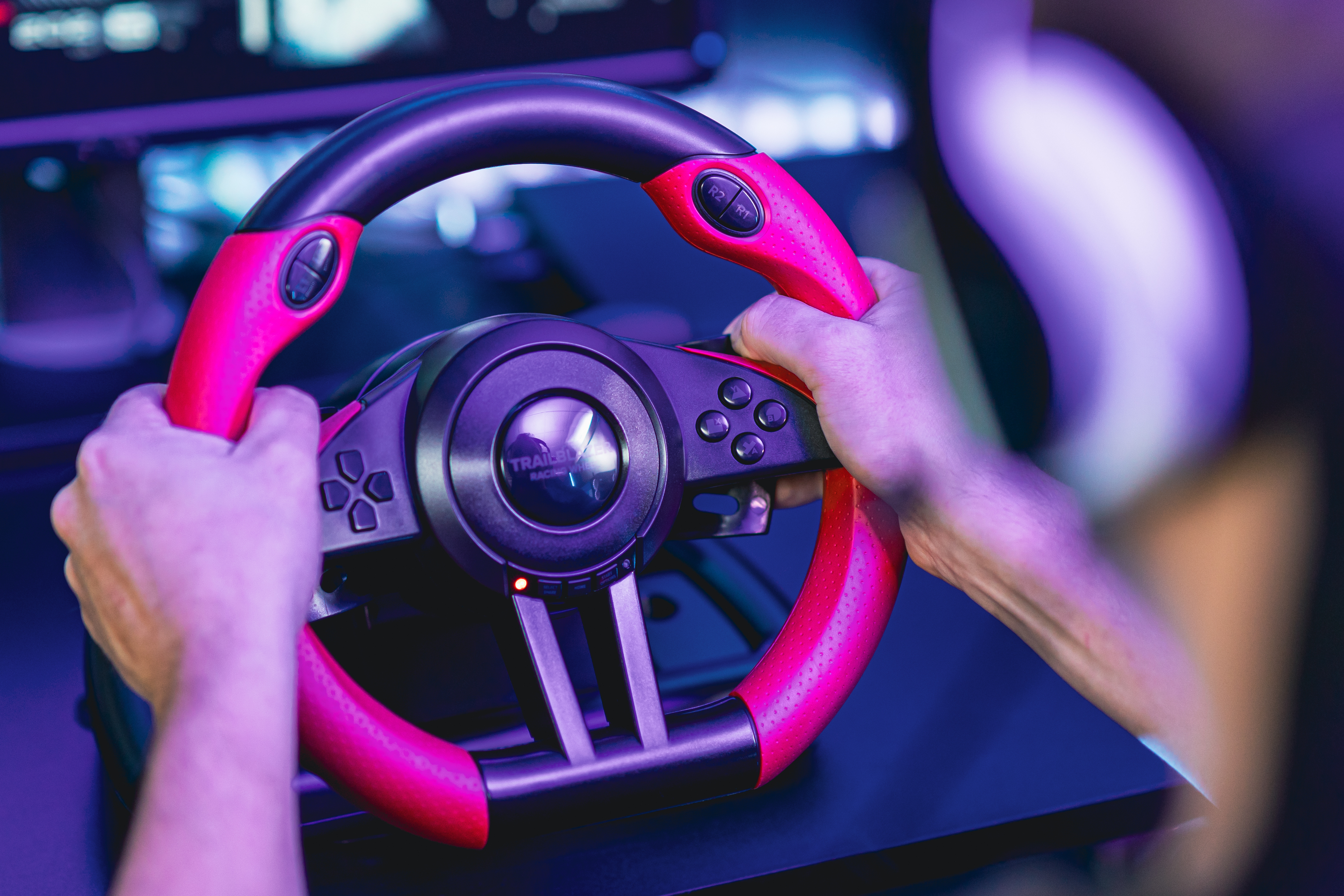 Hands gripping a pink and black racing wheel. Buttons and the Trailblazer logo are visible.