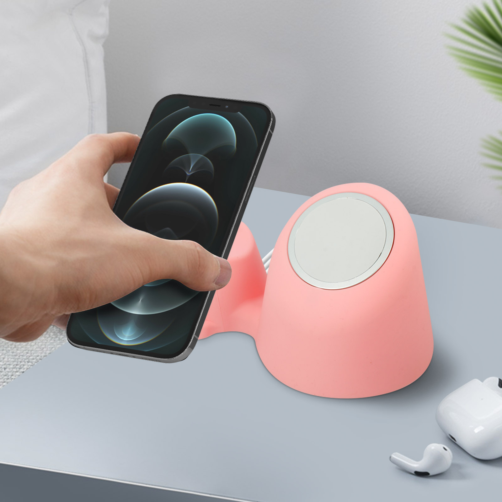 Una mano sostiene un teléfono sobre una estación de carga rosa, con AirPods en una superficie gris.