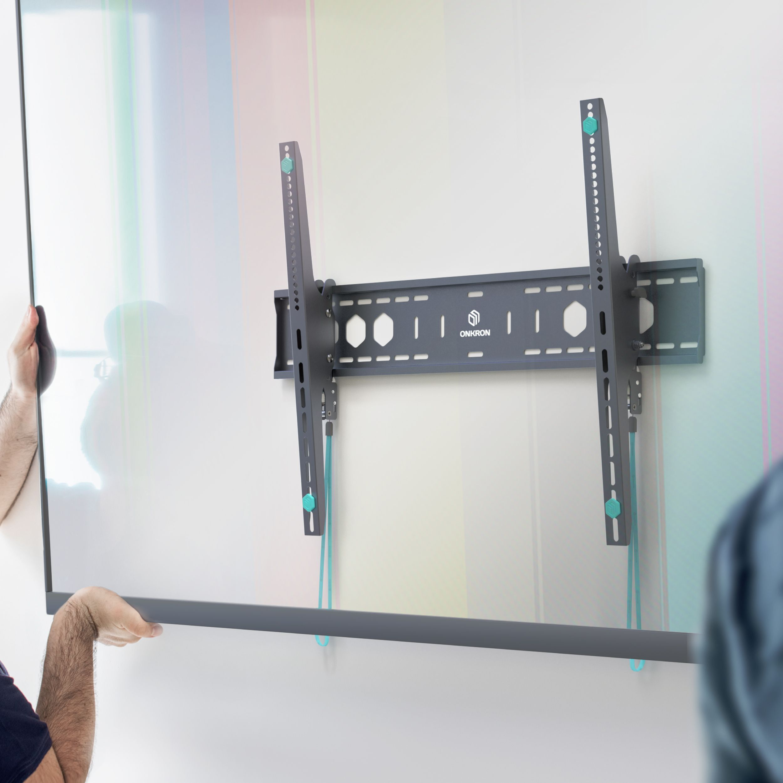 Osoba instaluje szary uchwyt do telewizora na białej ścianie. Uchwyt ma niebieskie akcenty. Druga osoba pomaga.