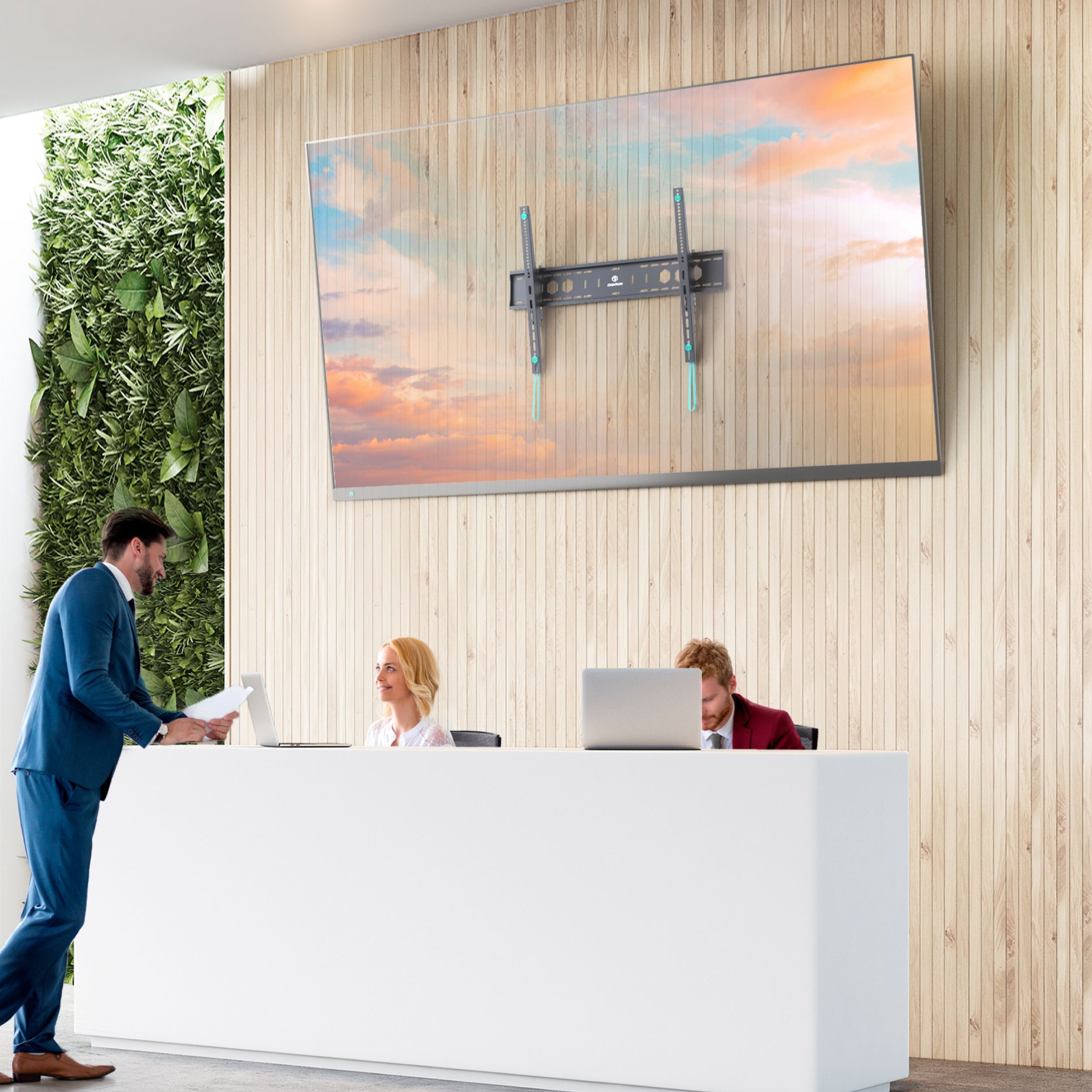 Recepcja biurowa z pracownikami, dużym telewizorem i ścianą roślin. Mężczyzna w niebieskim garniturze rozmawia z personelem.