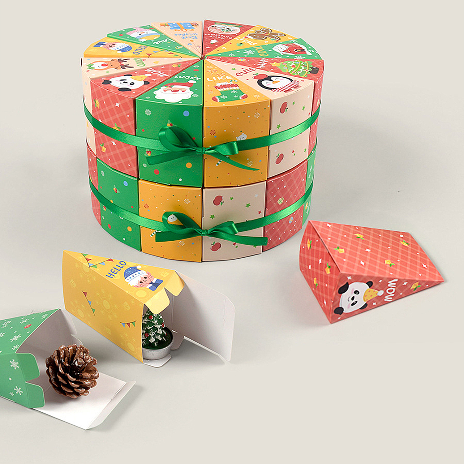Eine runde, geschichtete Geschenkbox mit grünem Band und mehrere offene dreieckige Schachteln mit Weihnachtsmotiven auf weißer Oberfläche.