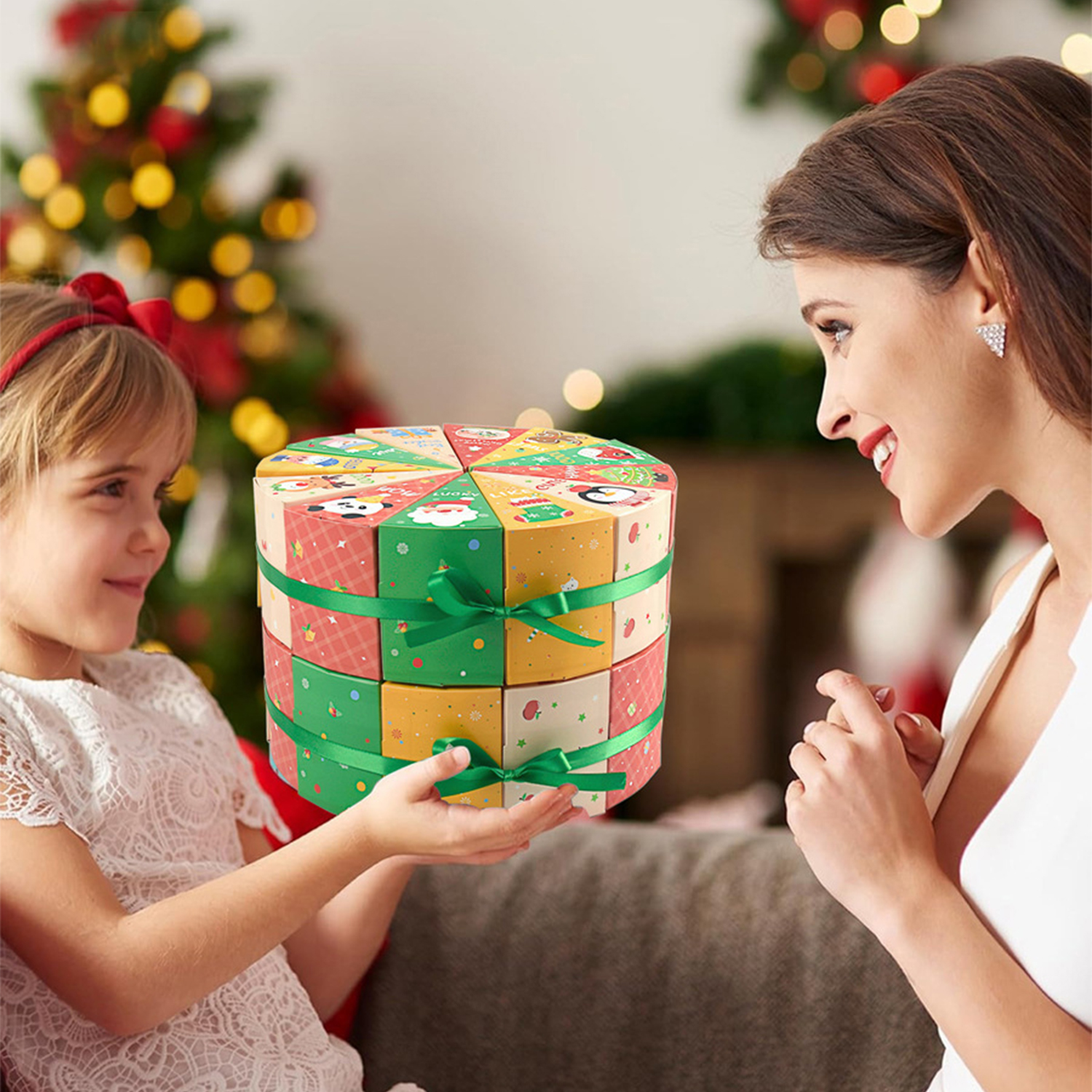 Ein Mädchen präsentiert einer Frau eine Weihnachtsgeschenkbox. Die Box ist bunt und mit grünen Bändern verpackt. Im Hintergrund ist ein Weihnachtsbaum.