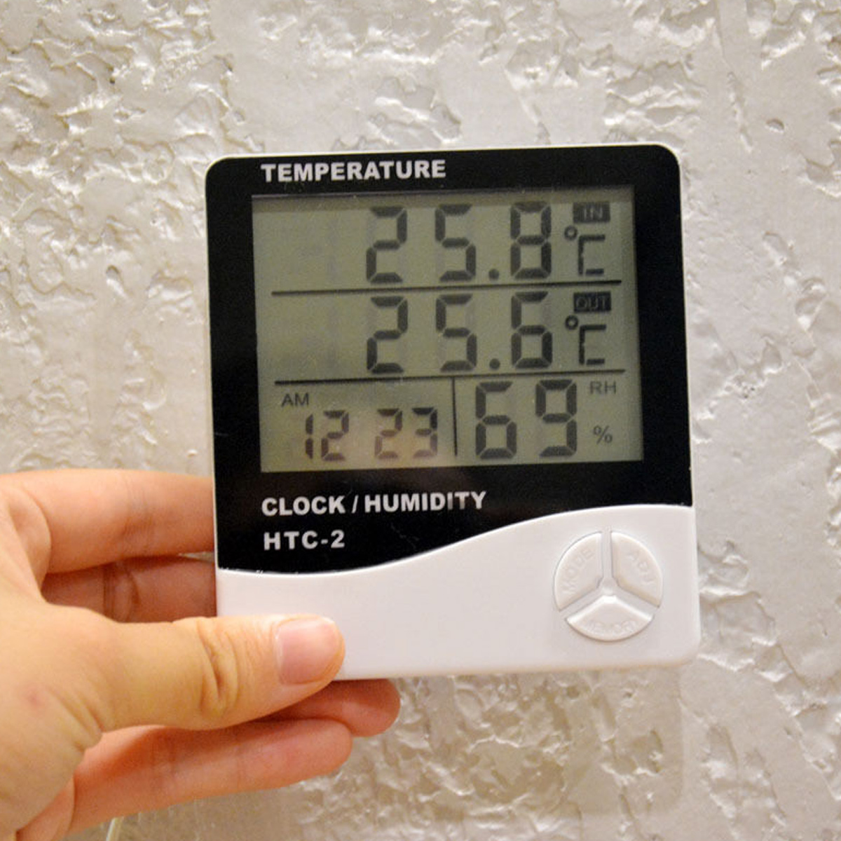 Eine Hand hält ein digitales Thermometer mit Temperaturen von 25,8°C und 25,6°C, Luftfeuchtigkeit 69% und Uhrzeit 12:23 Uhr. Hintergrund ist eine Wand.