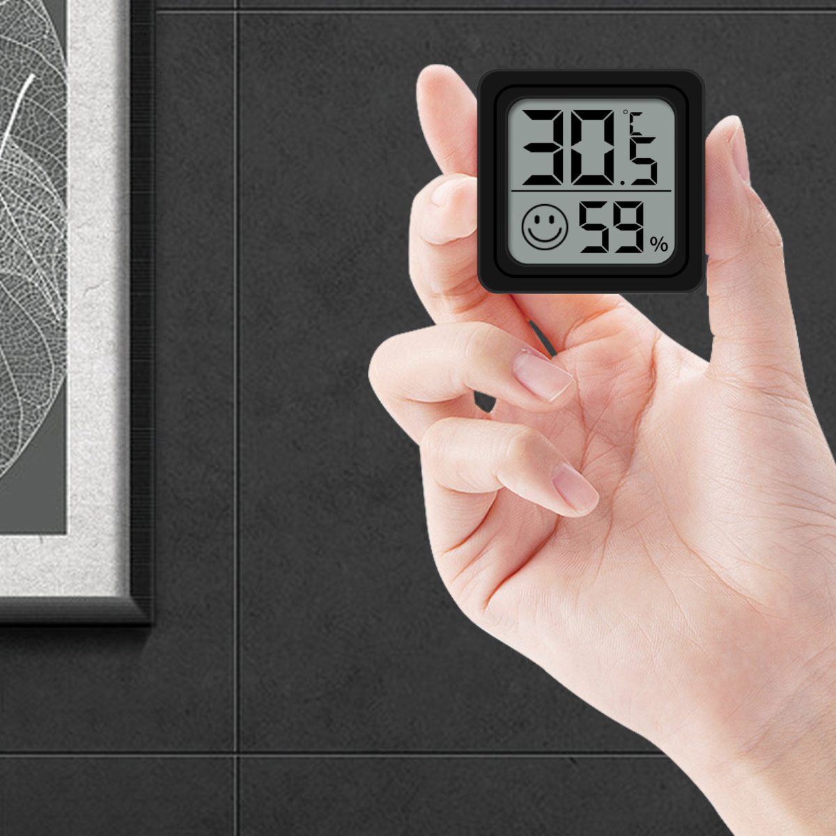 Eine Hand hält ein schwarzes digitales Thermometer, das 30,5 Grad und 59 % anzeigt. Im Hintergrund ist eine schwarze Wand und ein gerahmtes Bild zu sehen.