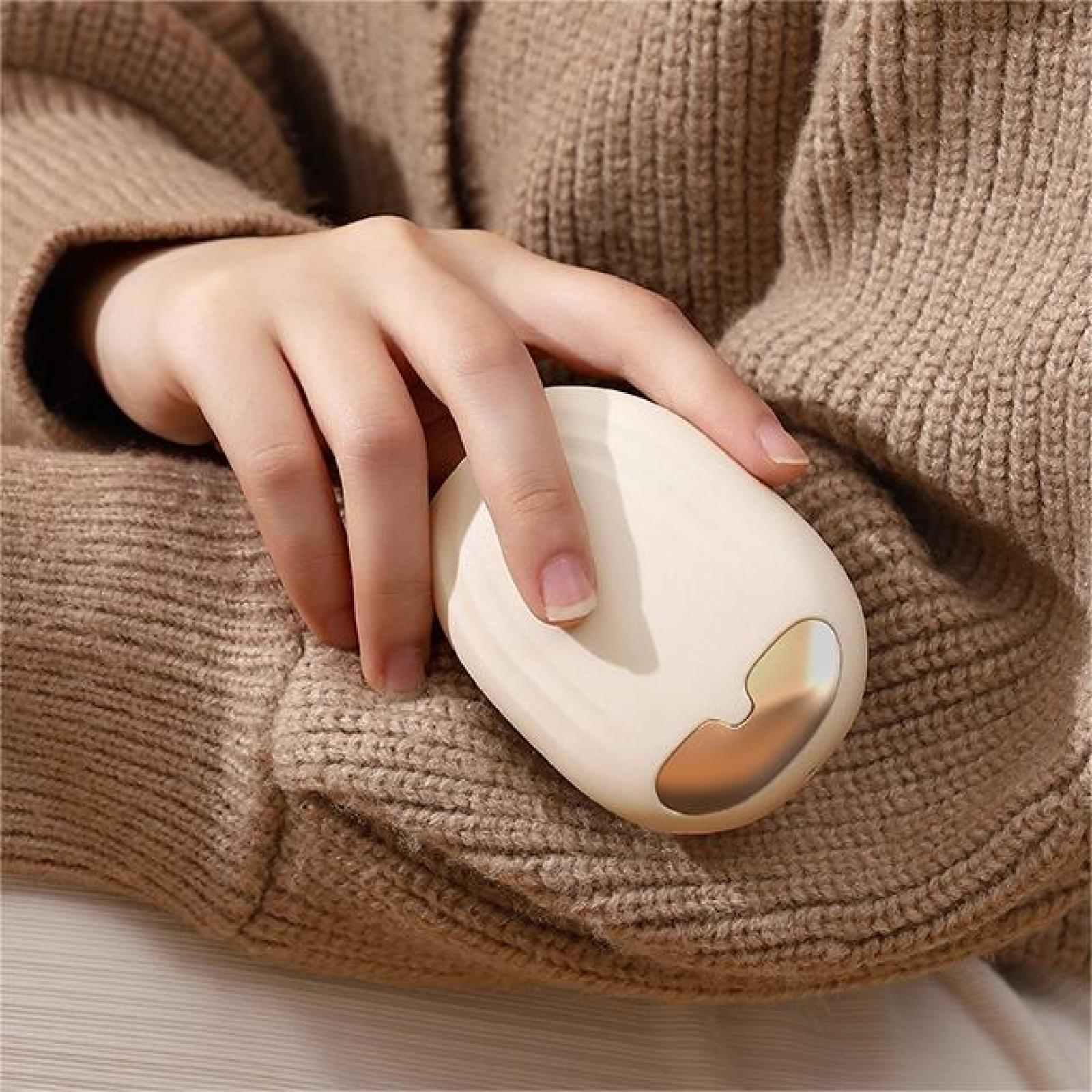 Eine Hand hält ein cremefarbenes, ovales Gerät mit goldenem Akzent. Die Person trägt einen braunen Strickpullover.