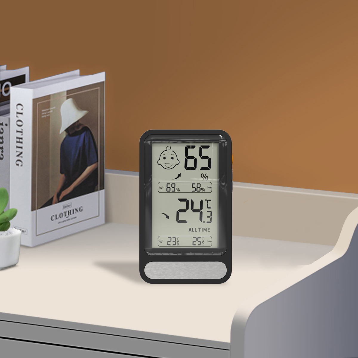 Ein digitales Thermometer zeigt Luftfeuchtigkeit und Temperatur an, platziert auf einer weißen Oberfläche. Bücher und eine kleine Pflanze sind im Hintergrund zu sehen.