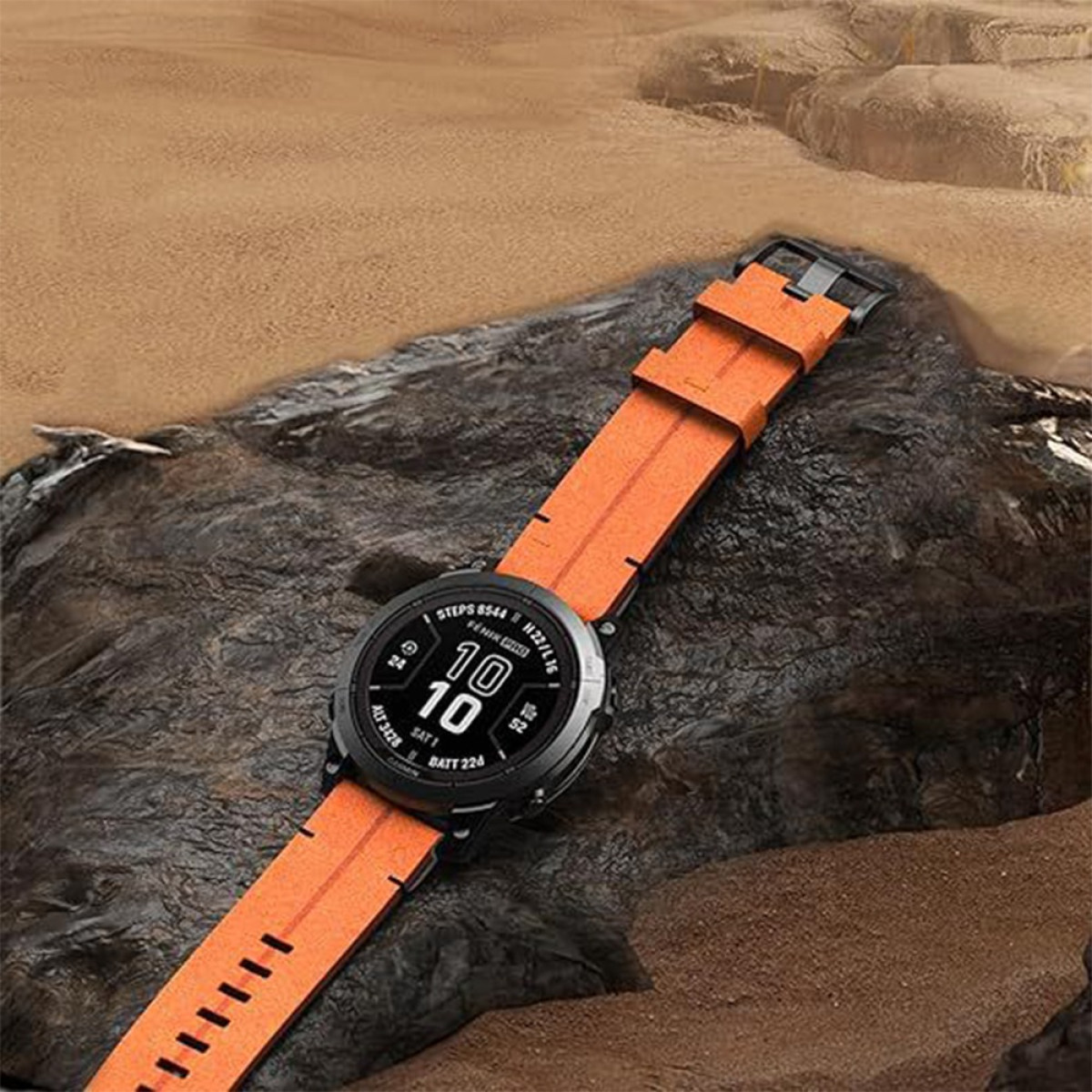 Eine schwarze Smartwatch mit orangefarbenem Armband liegt auf einem dunklen Felsen. Im Hintergrund sind Sand und Felsformationen zu sehen.