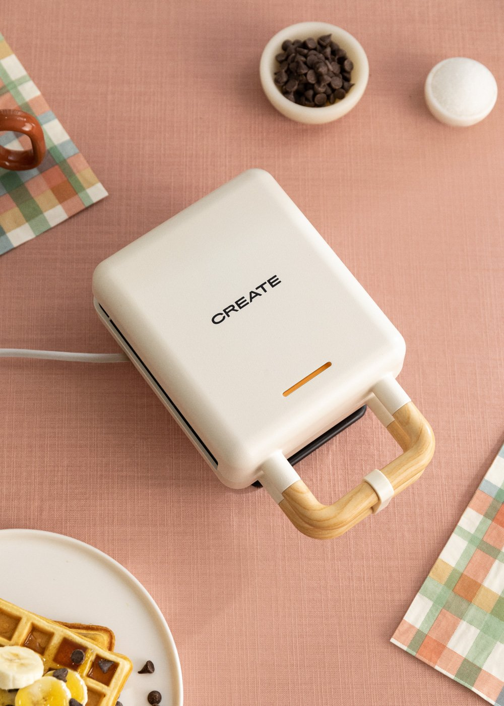 Una máquina para hacer gofres con 'CREATE' en la parte superior, con un plato de gofres y chispas de chocolate sobre una superficie rosa.