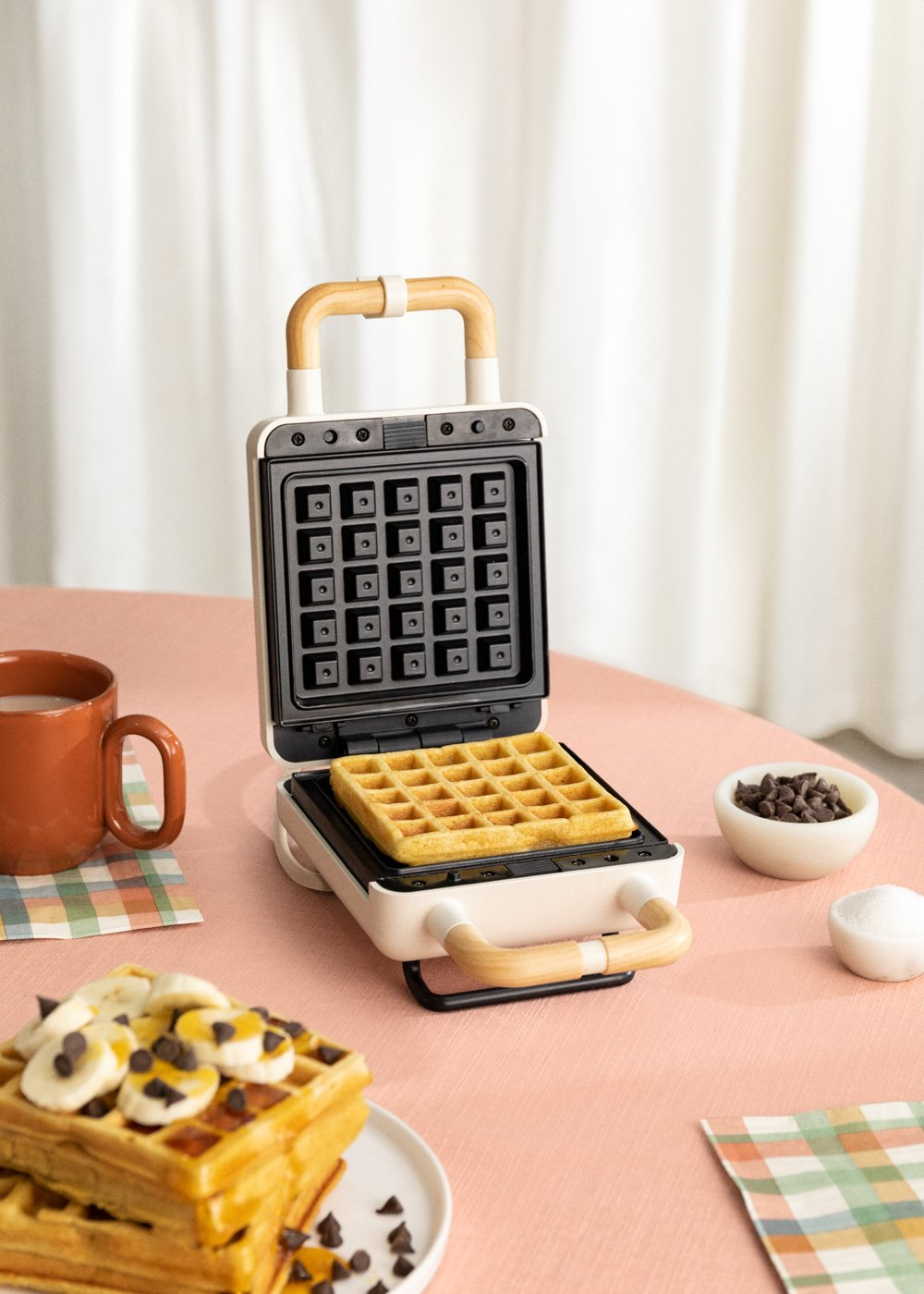 Una máquina para hacer gofres con un gofre cocido dentro. Los gofres están cubiertos con plátanos y chispas de chocolate, con café cerca.