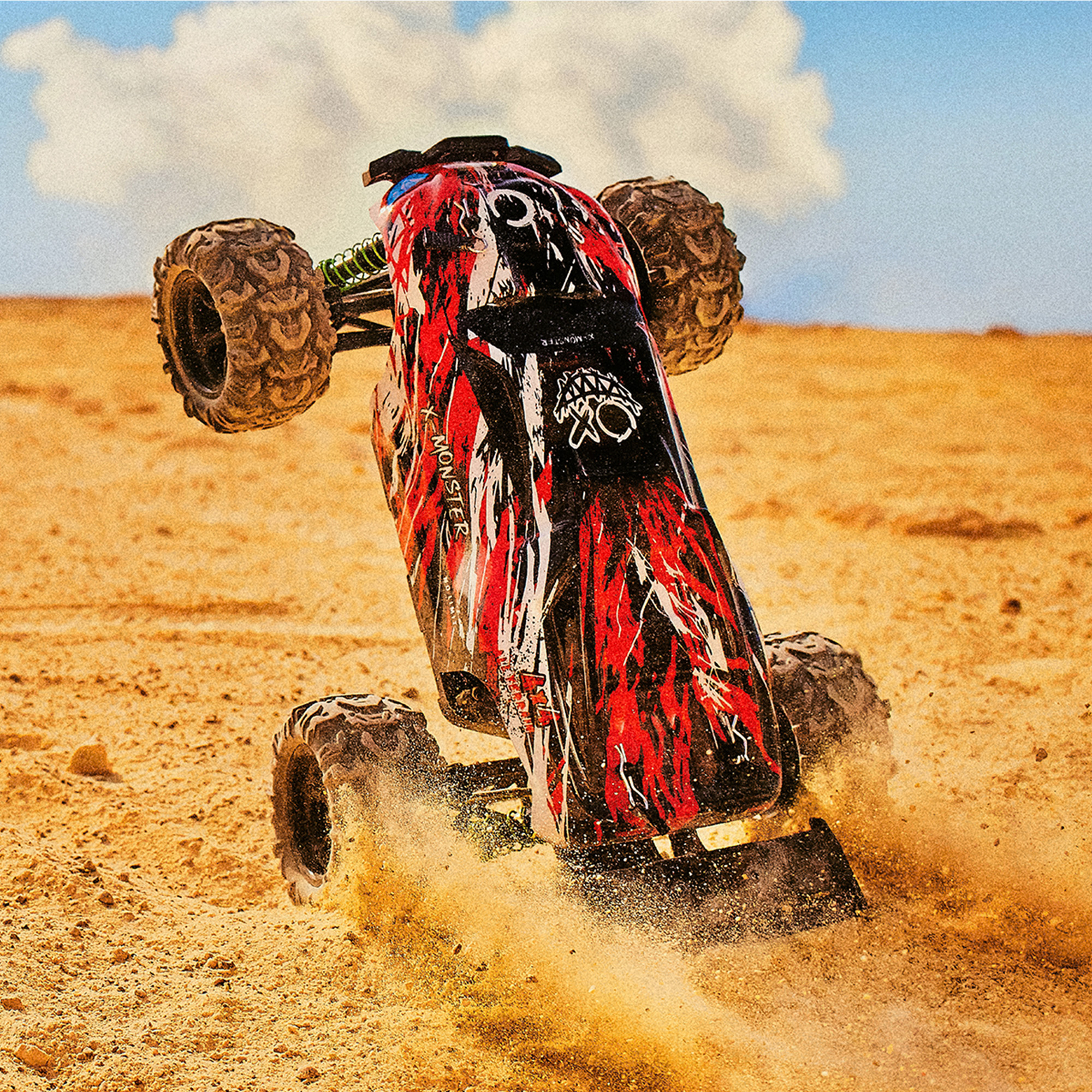 Czerwono-czarny monster truck RC jest w powietrzu na torze ziemnym, wzbijając kurz.