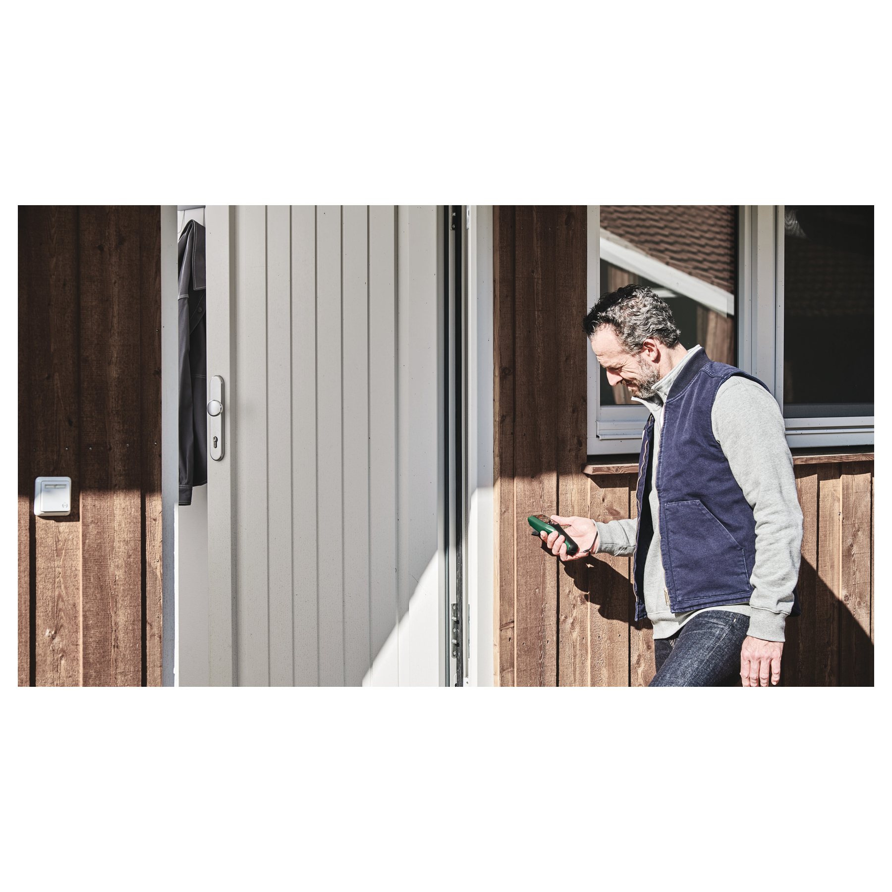 Homme aux cheveux gris, en gilet bleu, utilise un appareil vert. Près d'une porte blanche et d'un mur en bois.