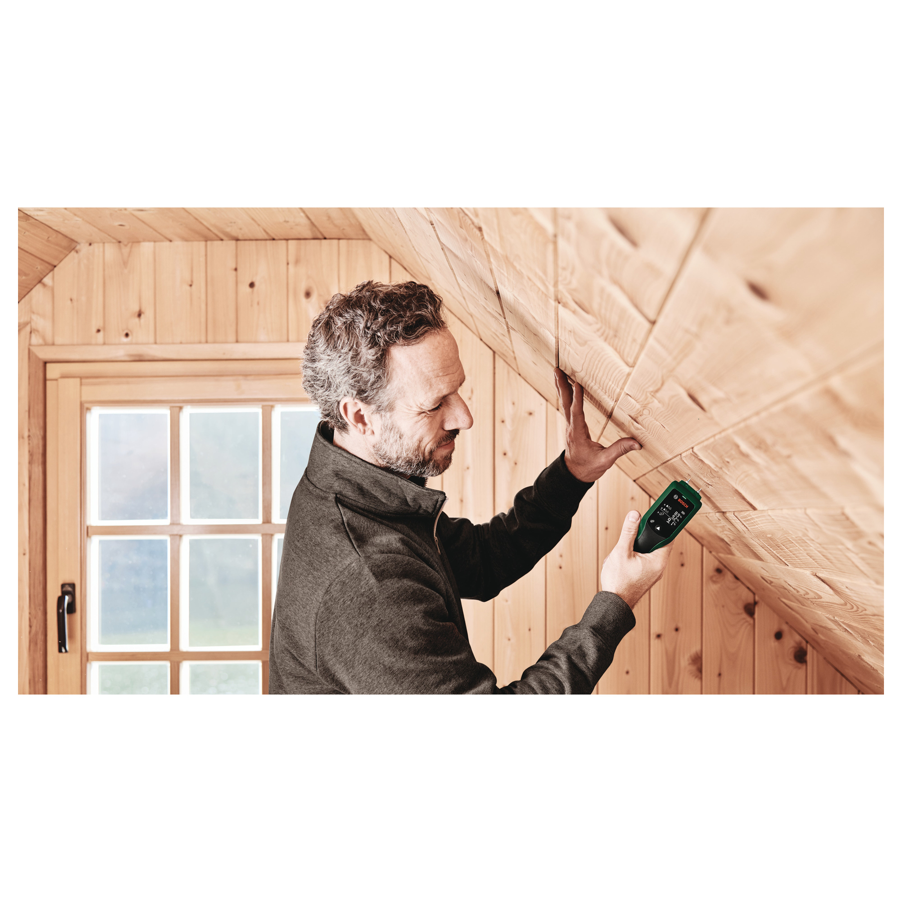 Homme utilisant un détecteur sur un plafond en bois. Il tient un appareil vert, évaluant le bois dans une cabane.
