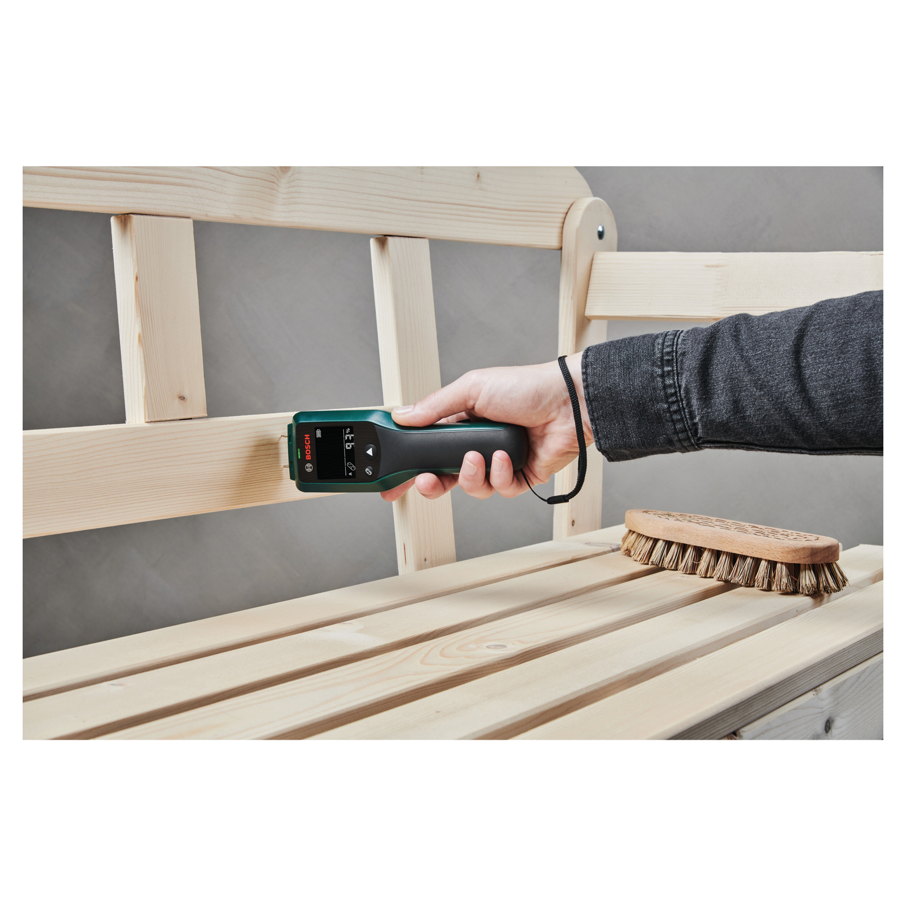 Une main tient un détecteur Bosch sur un banc en bois. Une brosse est à proximité. Le fond est gris.