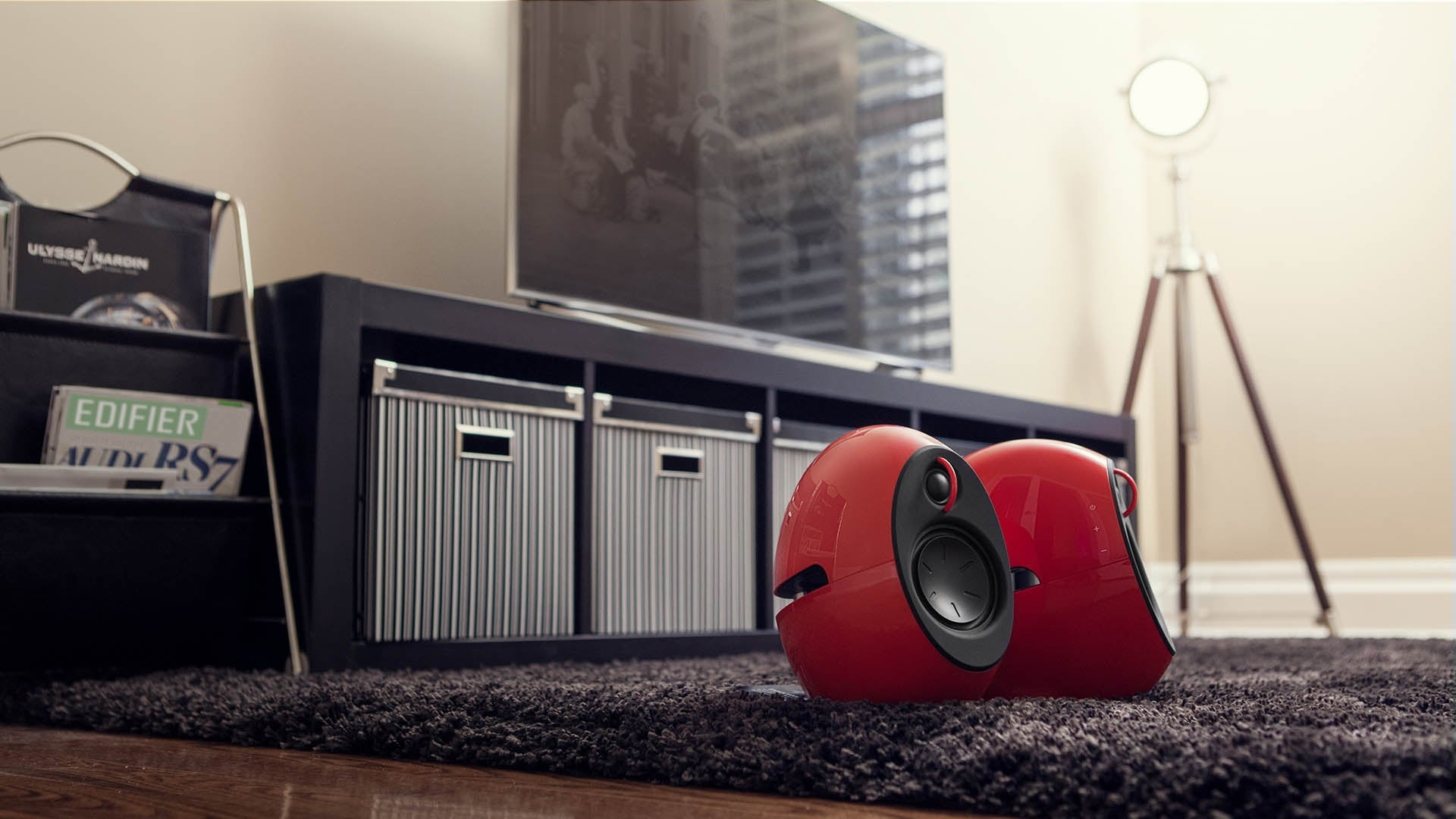 Czerwone głośniki na dywanie, z telewizorem i szafką w tle. Widoczna także lampa i książki.