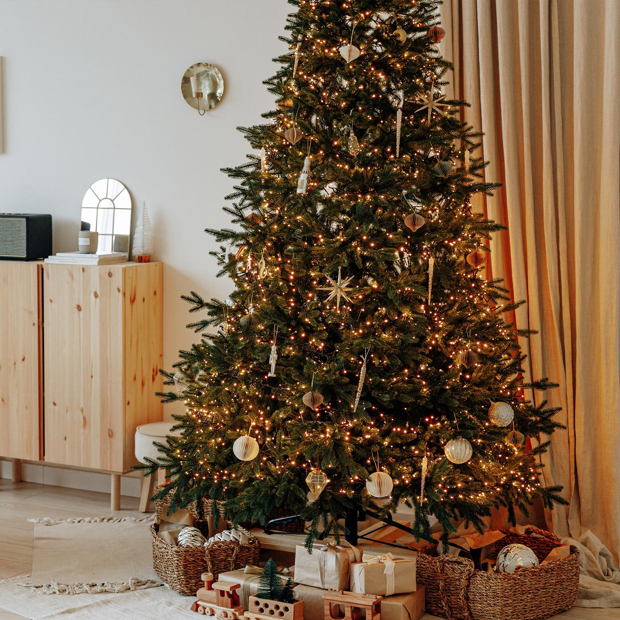 Weihnachtsbaum mit Ornamenten und Lichtern, Geschenke darunter. Weidenkörbe und ein Holzschrank sind im Raum.