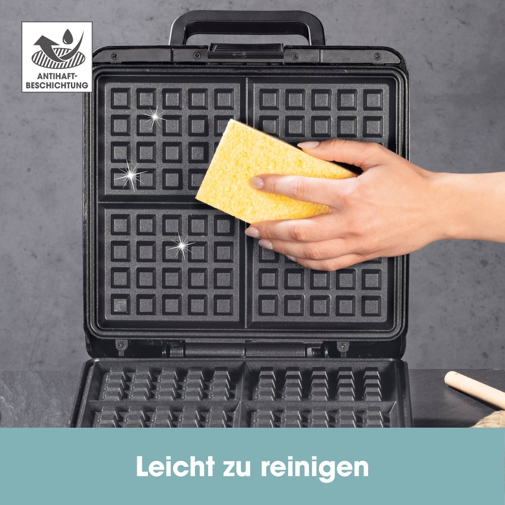 Eine Person reinigt ein Waffeleisen mit einem gelben Schwamm. Das Eisen ist schwarz und offen. Der Hintergrund ist eine graue Oberfläche.