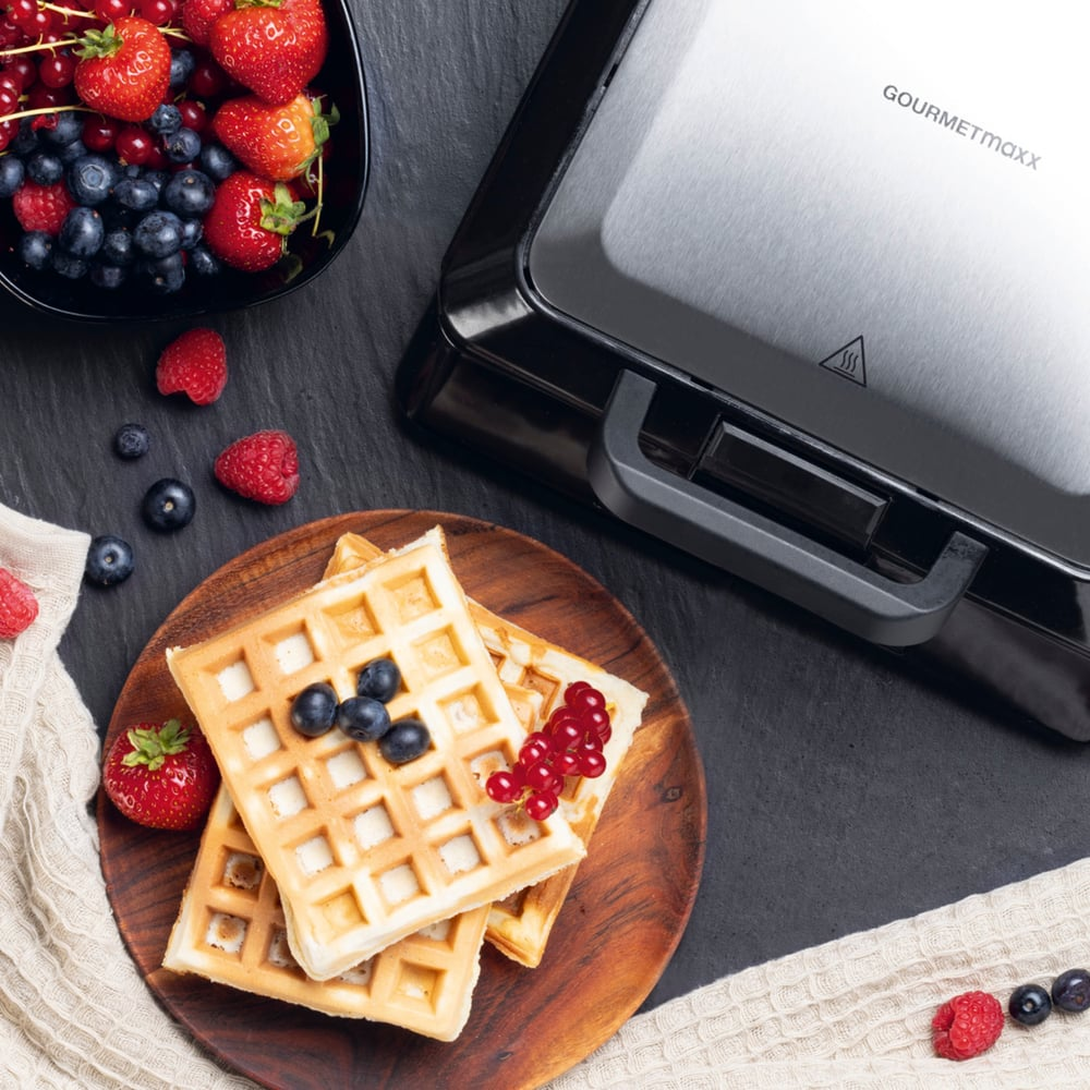 Waffeln auf Holzplatte mit Beeren und ein Waffeleisen auf schwarzer Oberfläche. Beeren in schwarzer Schüssel.
