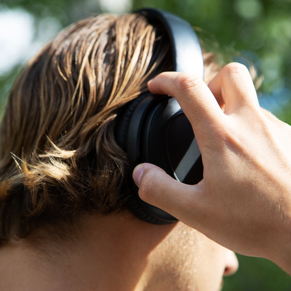 Osoba ma na sobie czarne słuchawki. Jej dłoń reguluje słuchawki. Tło jest rozmyte zielone.