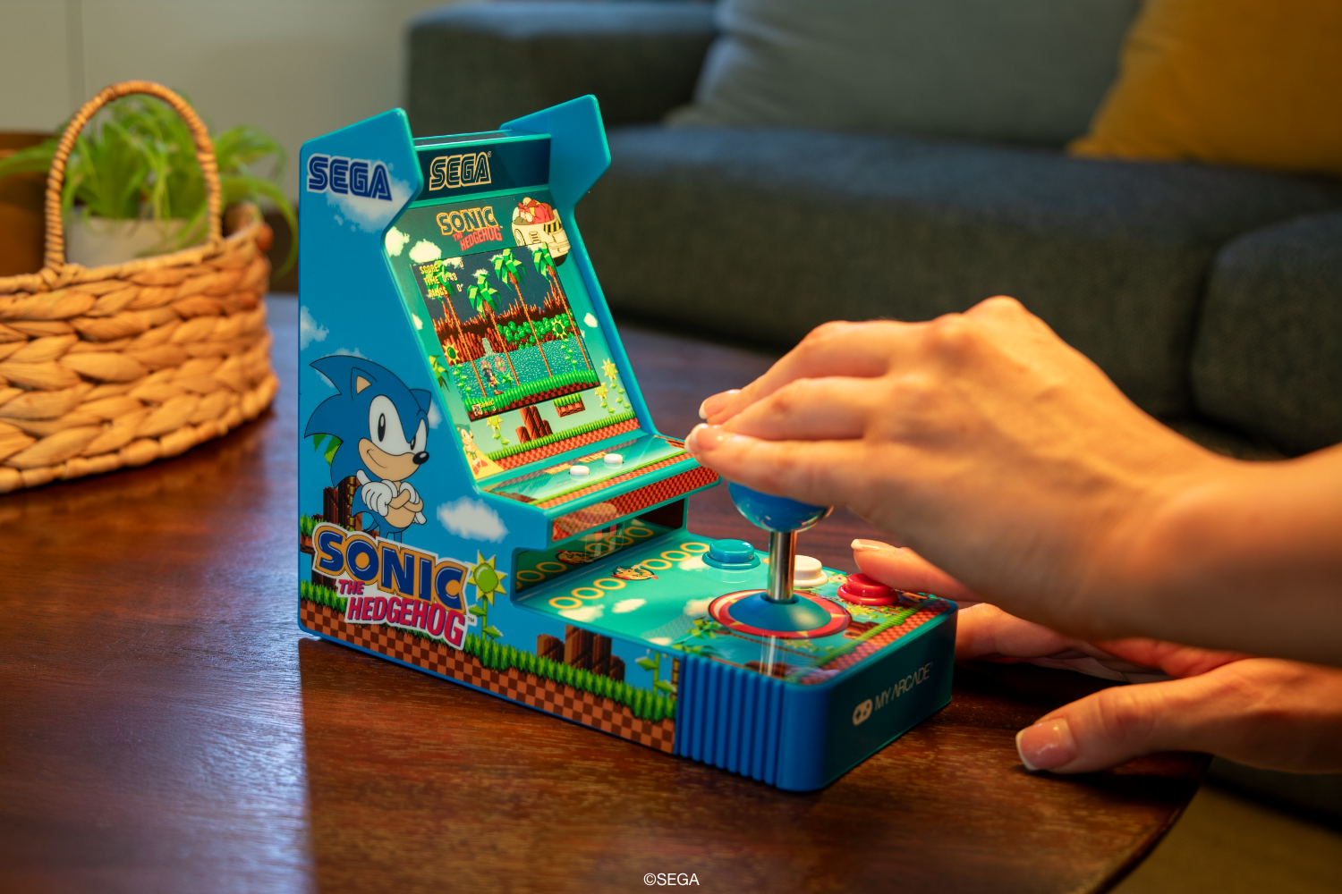 Eine Person spielt ein Sonic the Hedgehog Arcade-Spiel. Das Spiel ist blau. Eine Hand ist am Joystick. Im Hintergrund sind ein Korb und eine Couch.
