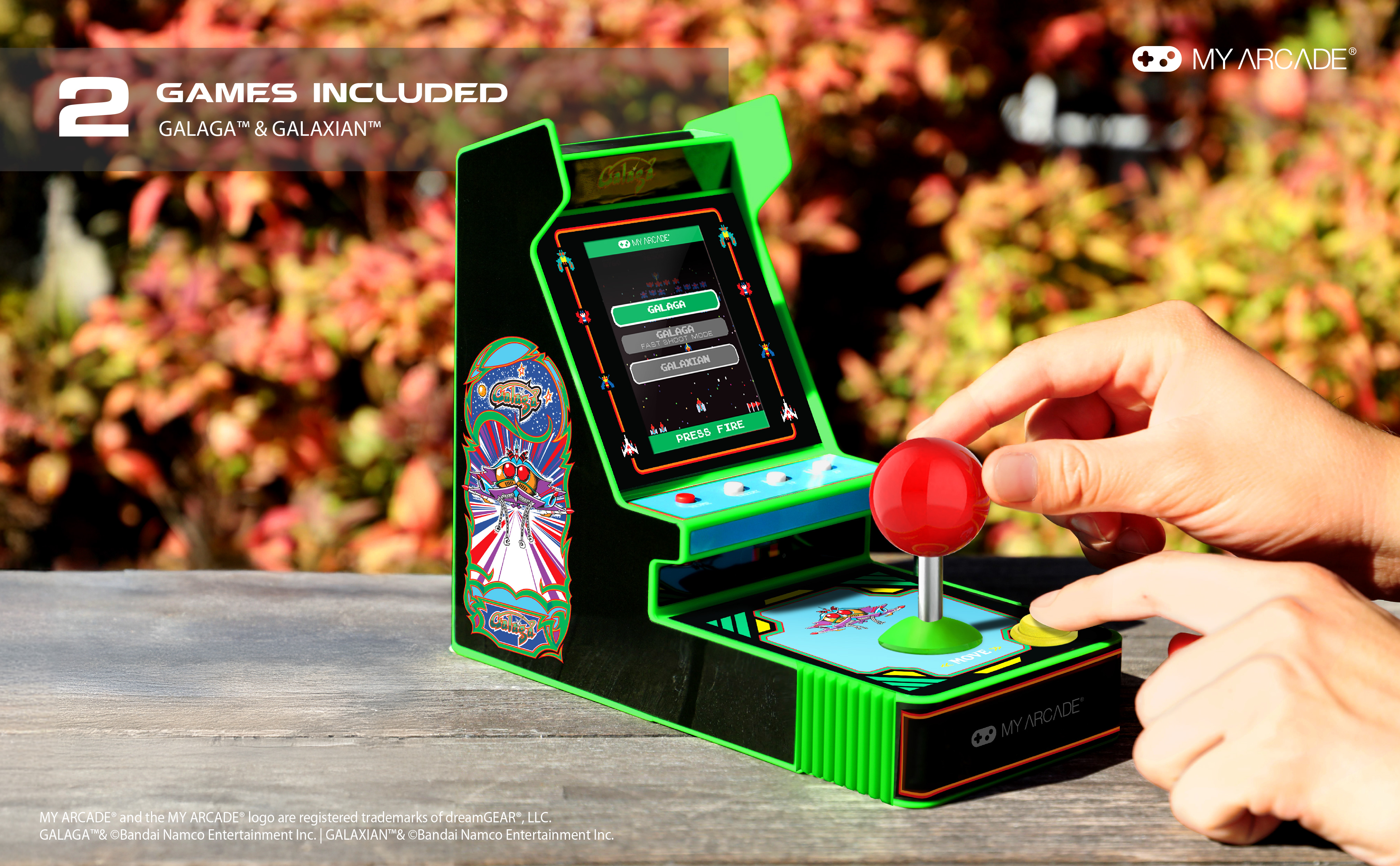 Mini cabinato arcade con joystick rosso, mostra Galaga e Galaxian. Mani visibili. Sfondo sfocato di foglie.