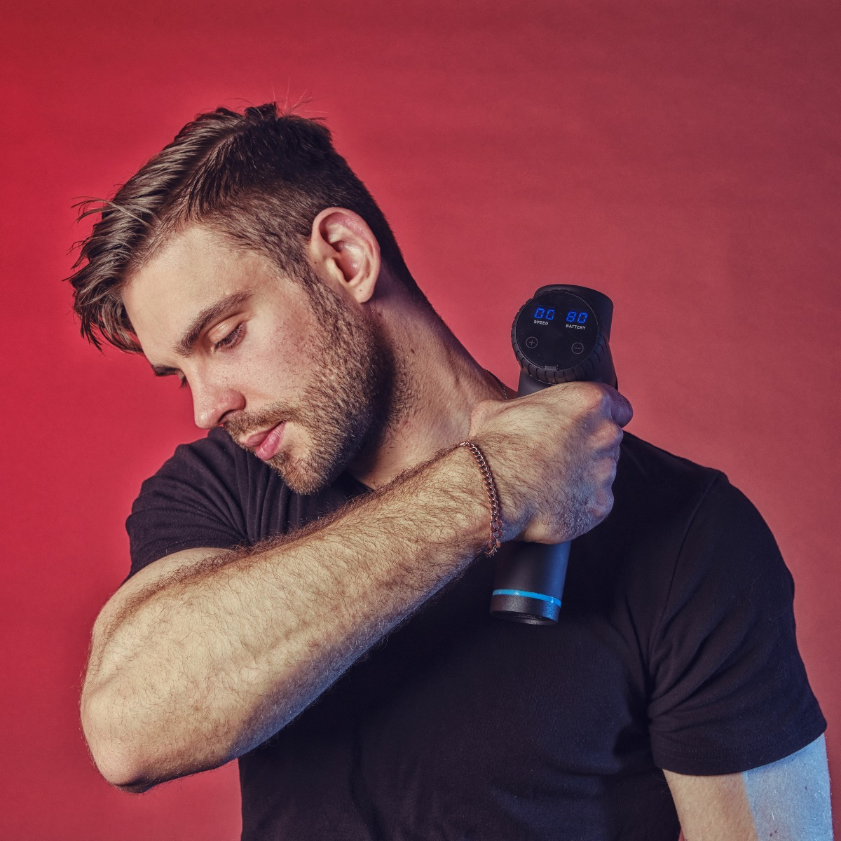 Mężczyzna w czarnej koszulce używa pistoletu do masażu na szyi na czerwonym tle. Patrzy w dół. Pistolet wyświetla prędkość i baterię.