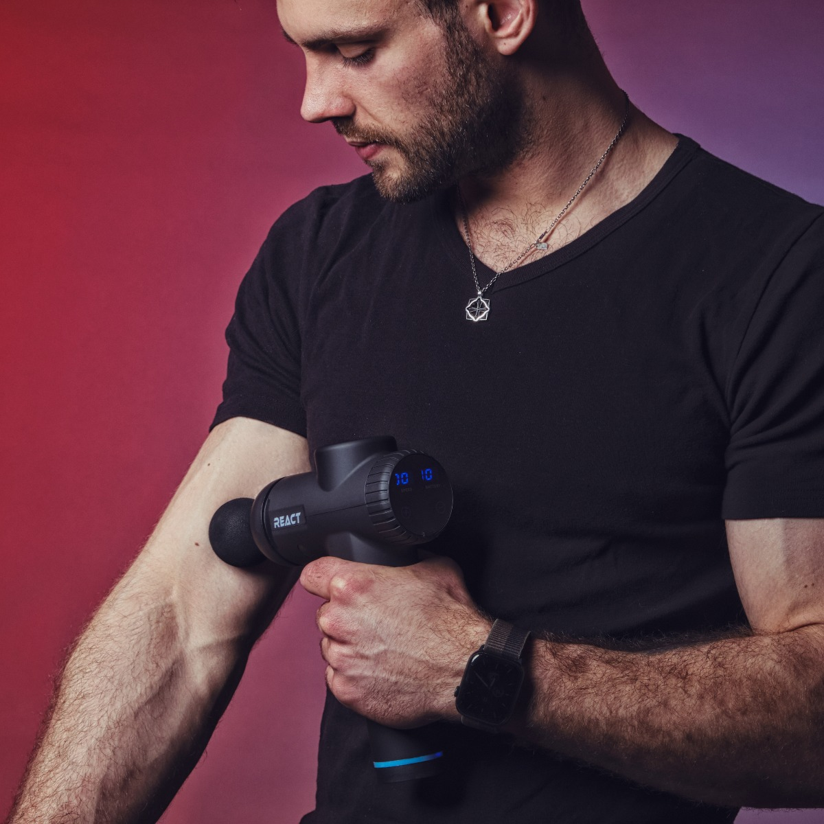 Mężczyzna w czarnej koszulce używa pistoletu do masażu na ramieniu. Nosi zegarek i naszyjnik. Tło jest czerwone i fioletowe.