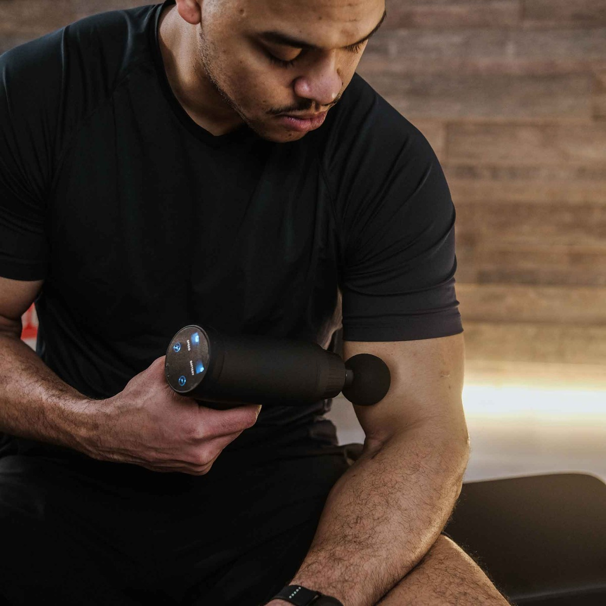 Mężczyzna używa czarnego pistoletu do masażu na ramieniu. Ma na sobie czarną koszulę, a tło to drewniana ściana.