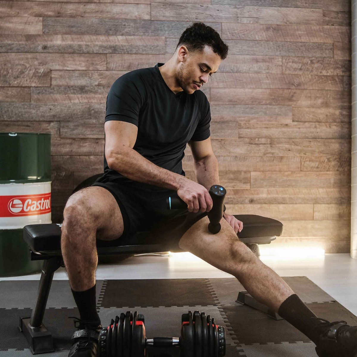 Mężczyzna w czerni używa pistoletu do masażu na nodze. Siedzi na ławce, w tle hantle i beczka.