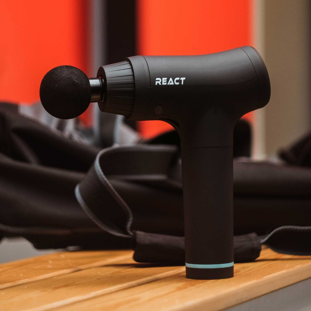 Czarny pistolet do masażu React na drewnianej powierzchni, z rozmytą czarną torbą w tle na pomarańczowej ścianie.