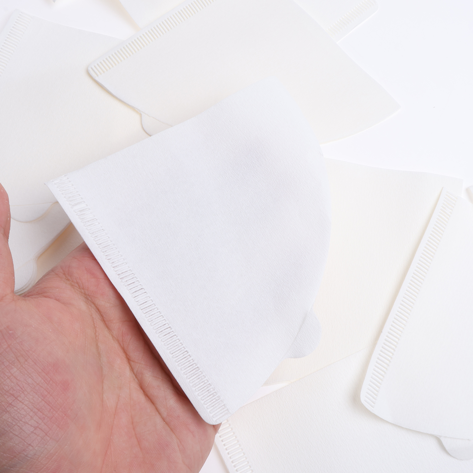 Eine Hand hält einen weißen Papier-Kaffeefilter. Weitere Filter liegen im Hintergrund auf weißem Untergrund verstreut.