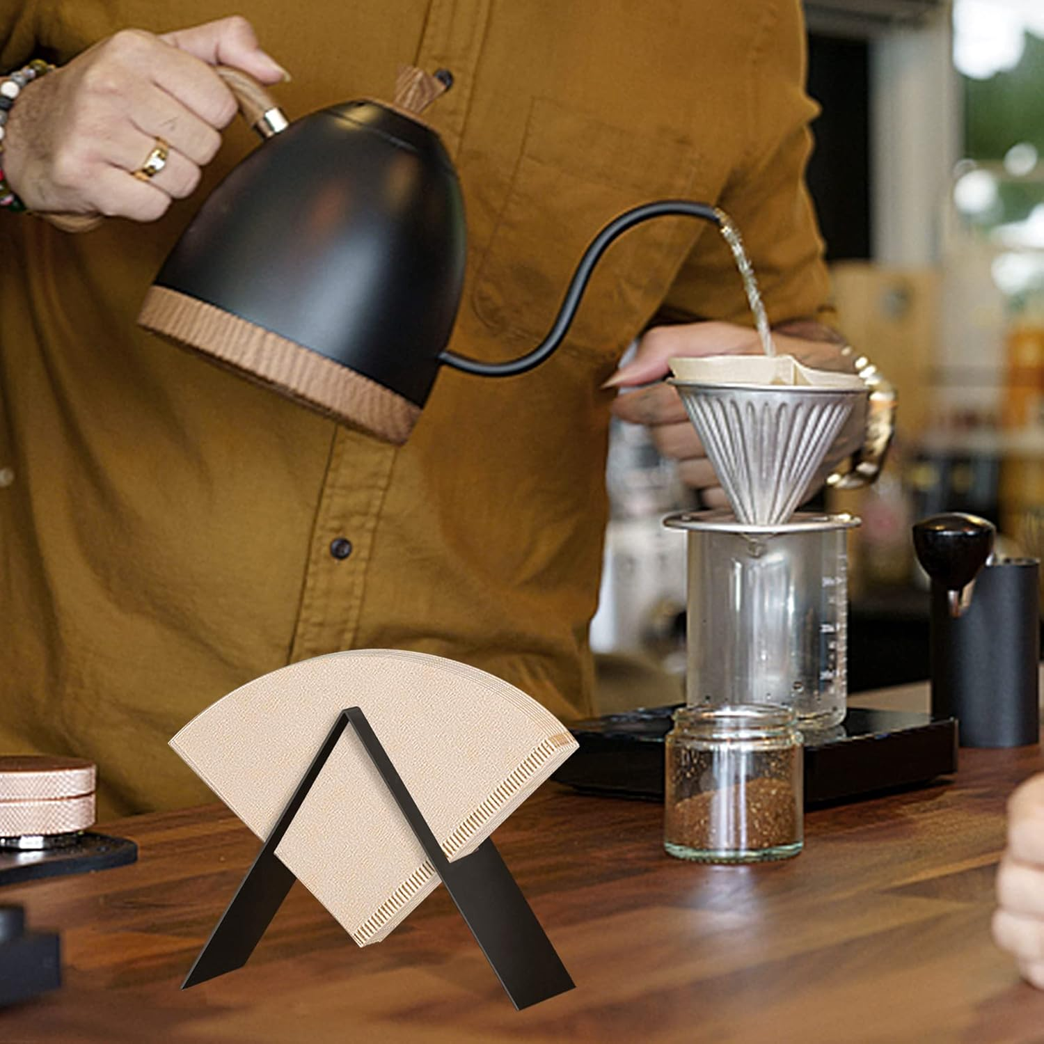 Eine Person gießt Wasser aus einem schwarzen Wasserkocher in einen Kaffeefilter. Kaffeefilter und andere Brühutensilien stehen auf Holz.