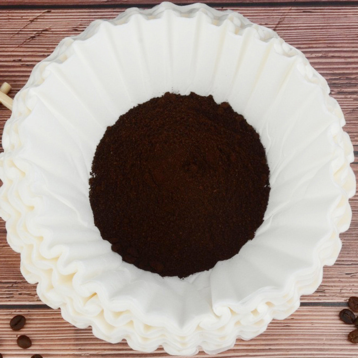 Ein weißer Kaffeefilter gefüllt mit dunkelbraunem Kaffeepulver liegt auf einer Holzoberfläche. Kaffeebohnen sind verstreut.