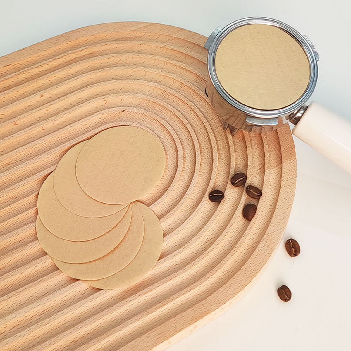 Ein Holztablett mit Kaffeefiltern und -bohnen. Rechts befindet sich ein Kaffeefilterträger. Weiße Oberfläche.