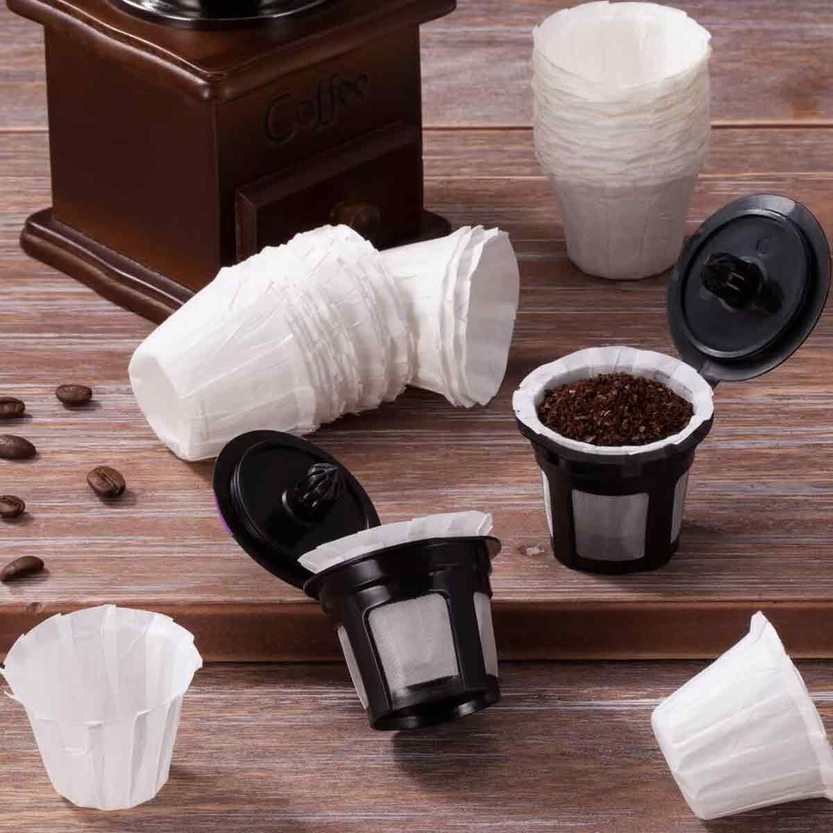 Kaffeepads, Filter und eine Kaffeemühle auf einem Holztisch. Einige Pads sind offen und mit Kaffeepulver gefüllt. Weiße Papierbecher vorhanden.