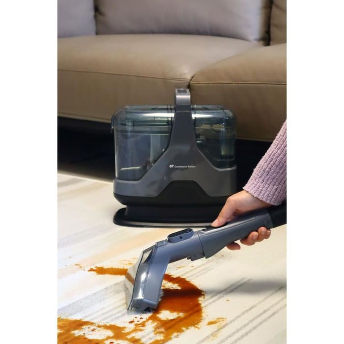 Eine Person reinigt mit einem grauen Teppichreiniger einen befleckten Teppich. Der Reiniger steht in der Nähe eines beigen Sofas.