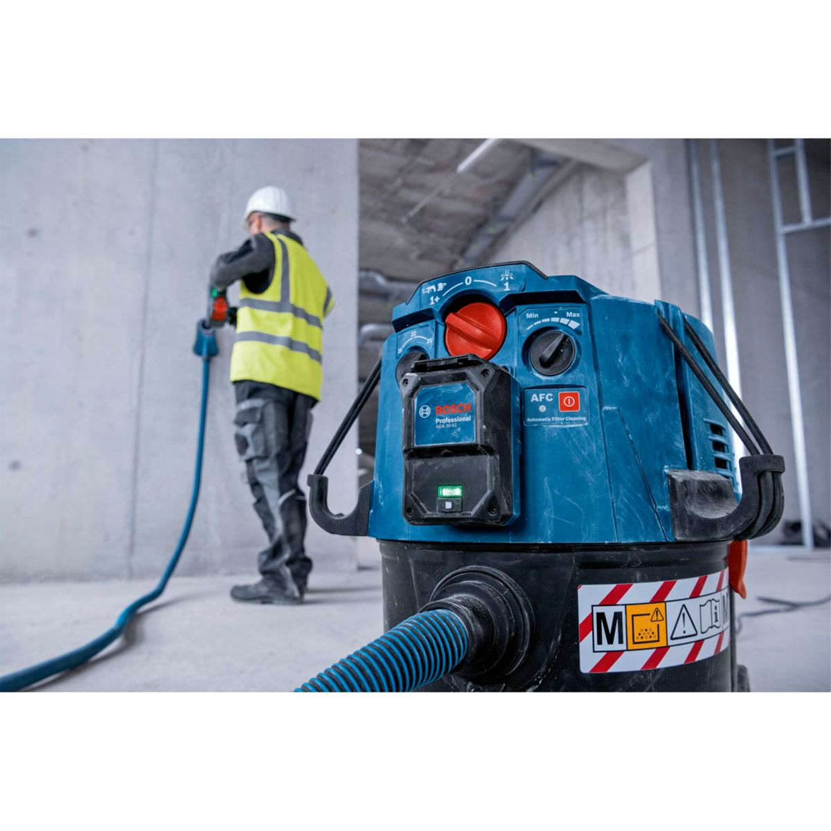 Un trabajador de la construcción usa una manguera azul para limpiar con una aspiradora azul y negra. El trabajador usa un chaleco amarillo y casco.