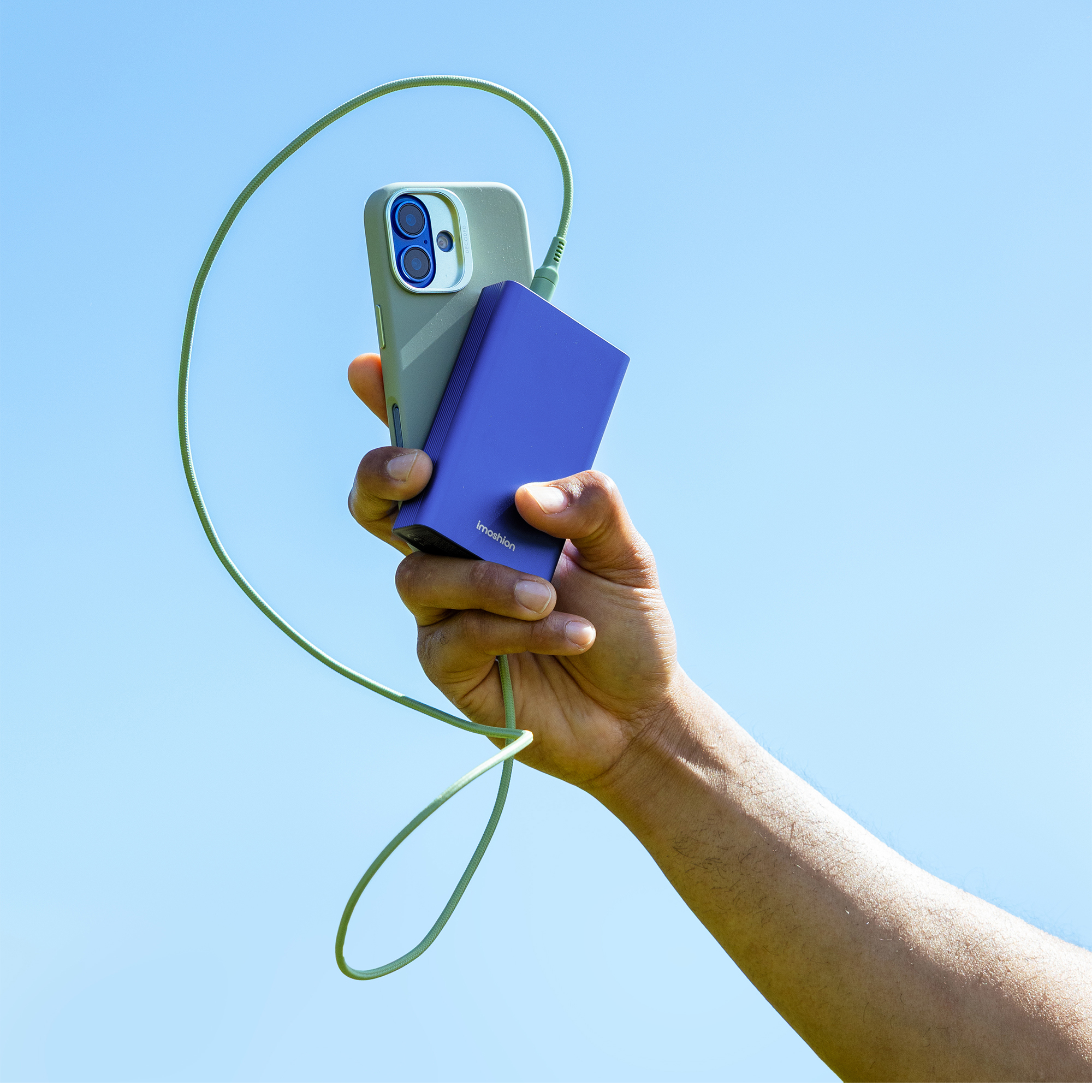 Main tenant téléphone et chargeur avec câble vert contre ciel bleu. Le chargeur est violet.