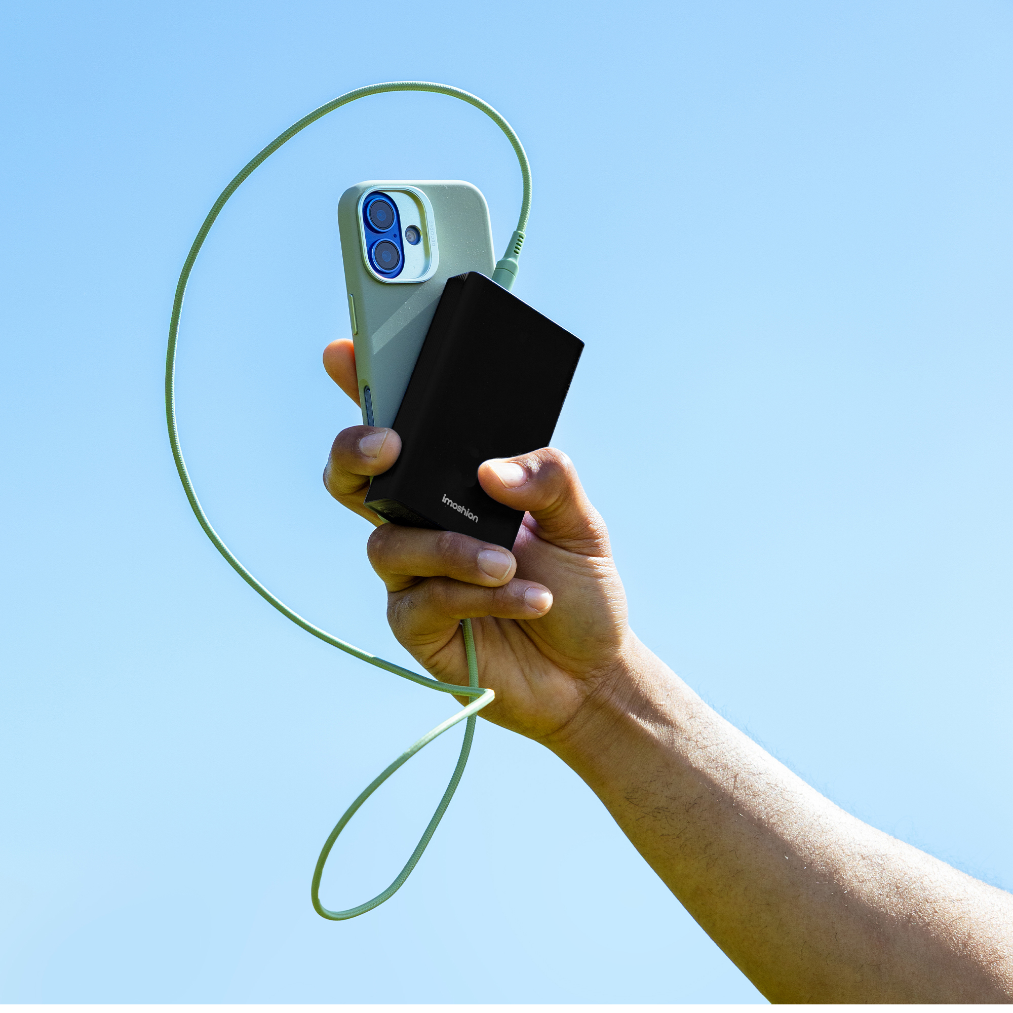 Main tenant un téléphone et une batterie externe noire avec un câble vert sur ciel bleu.