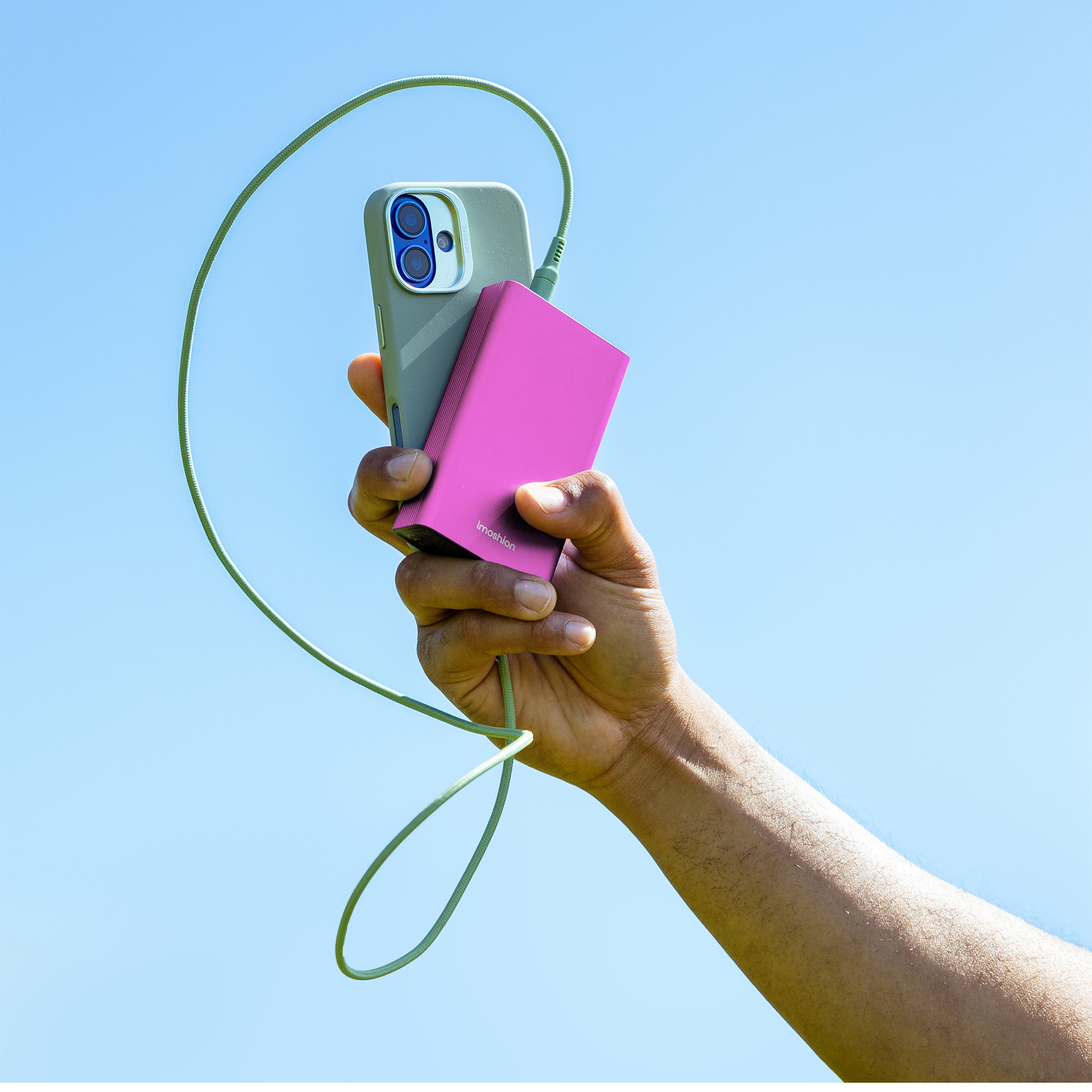 Une main tient un téléphone et une batterie externe rose. Un câble vert les relie. Ciel bleu en arrière-plan.
