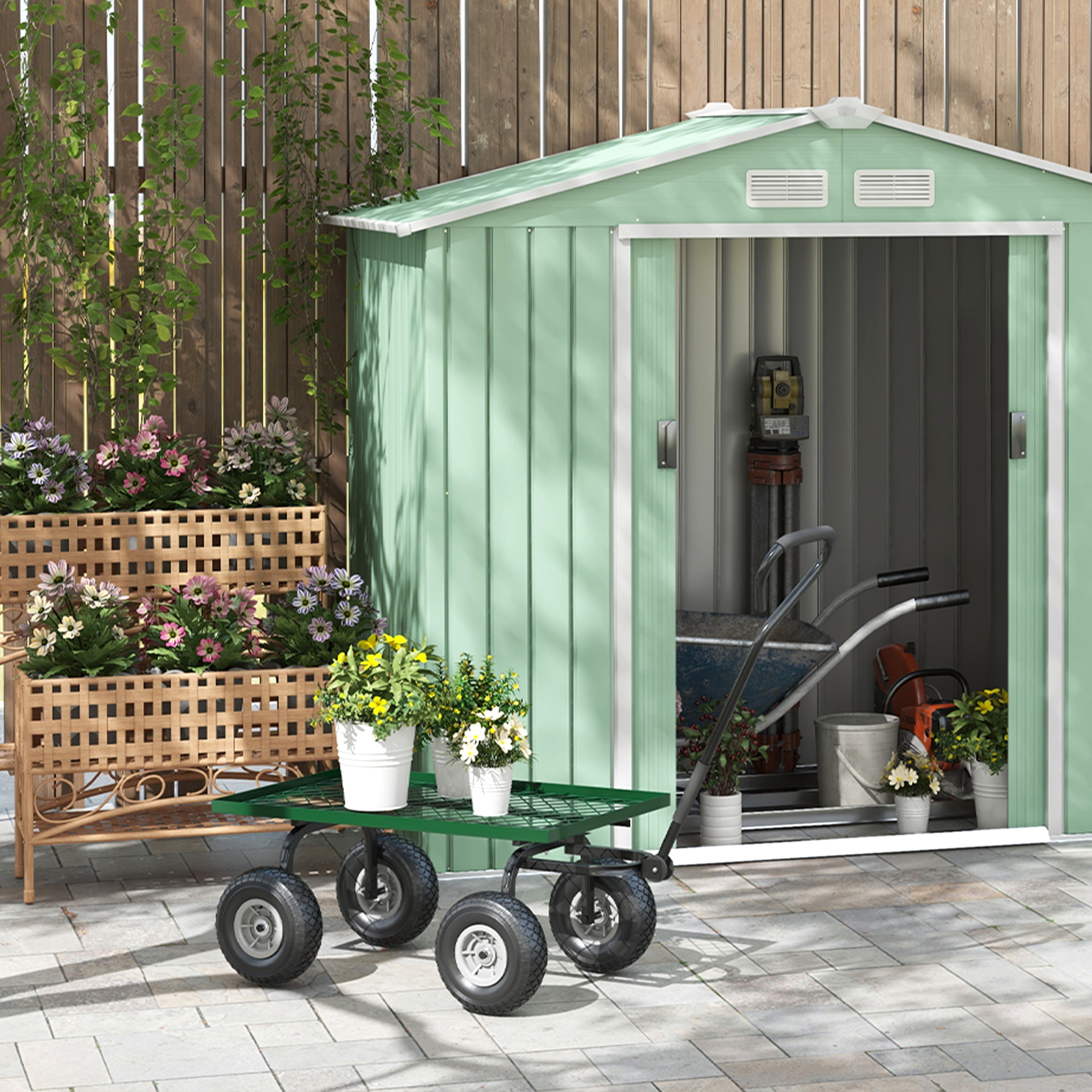 Un capannone verde con la porta aperta. Un carrello con le ruote è davanti. Fiori in primo e secondo piano.