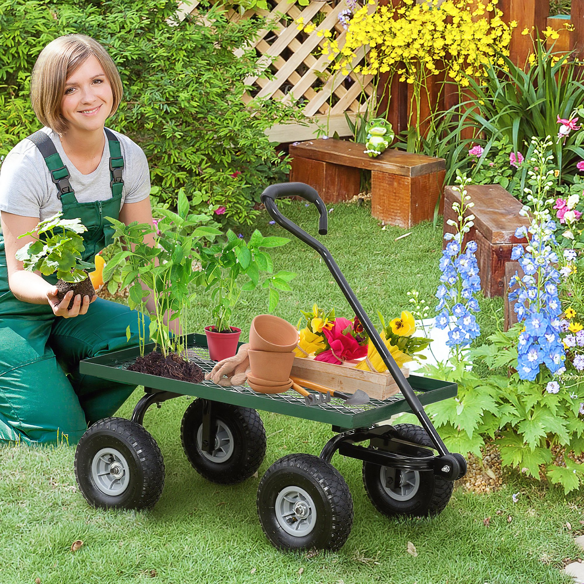 Donna in tuta che si inginocchia vicino a un carrello verde pieno di piante, vasi e attrezzi, in un giardino fiorito.