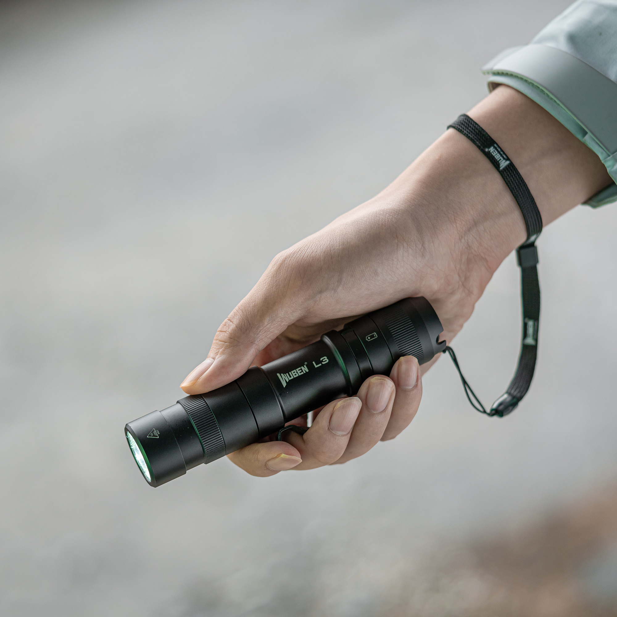 Eine Hand hält eine schwarze Taschenlampe, die Wuben L3, mit einer Handschlaufe. Der Hintergrund ist unscharf grau.