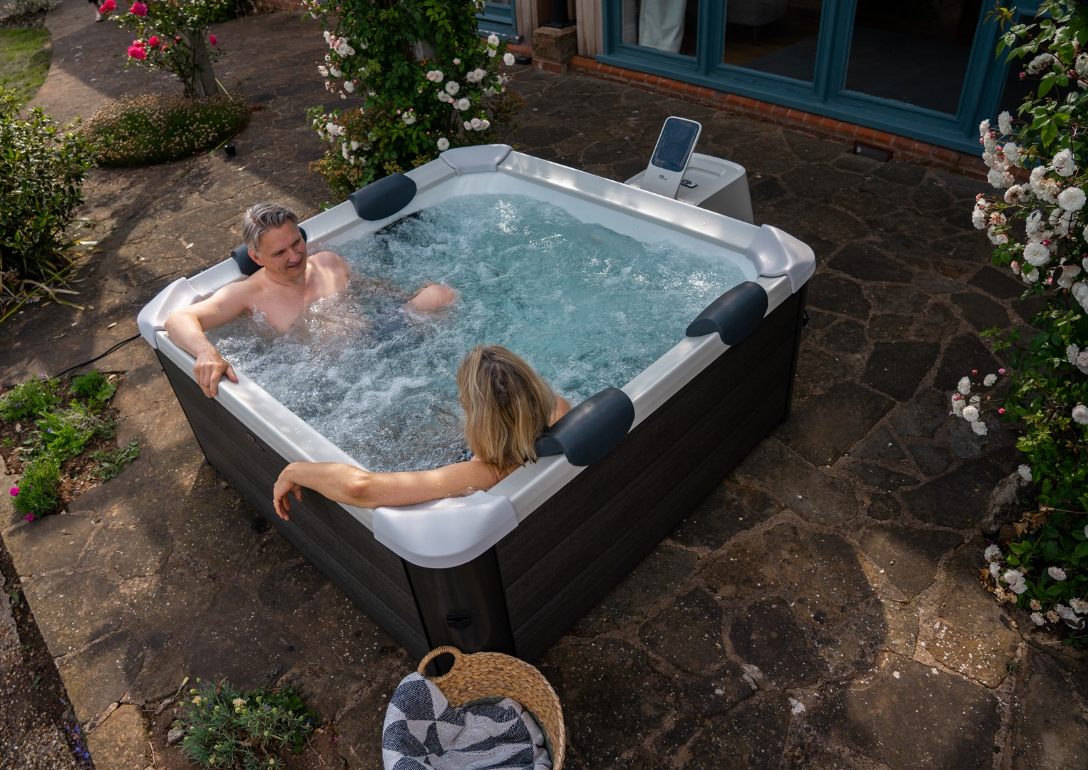 Ein Paar entspannt sich im Whirlpool. Die Wanne ist von Stein, Blumen und Pflanzen umgeben. Eine Person ist blond, die andere grauhaarig.
